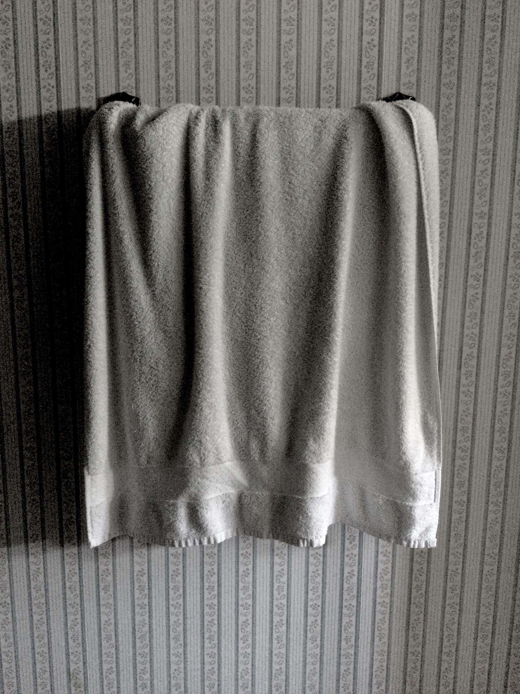 A towel hangs on a rack, many folds catching light from the right, with stripy old-timey wallpaper behind.