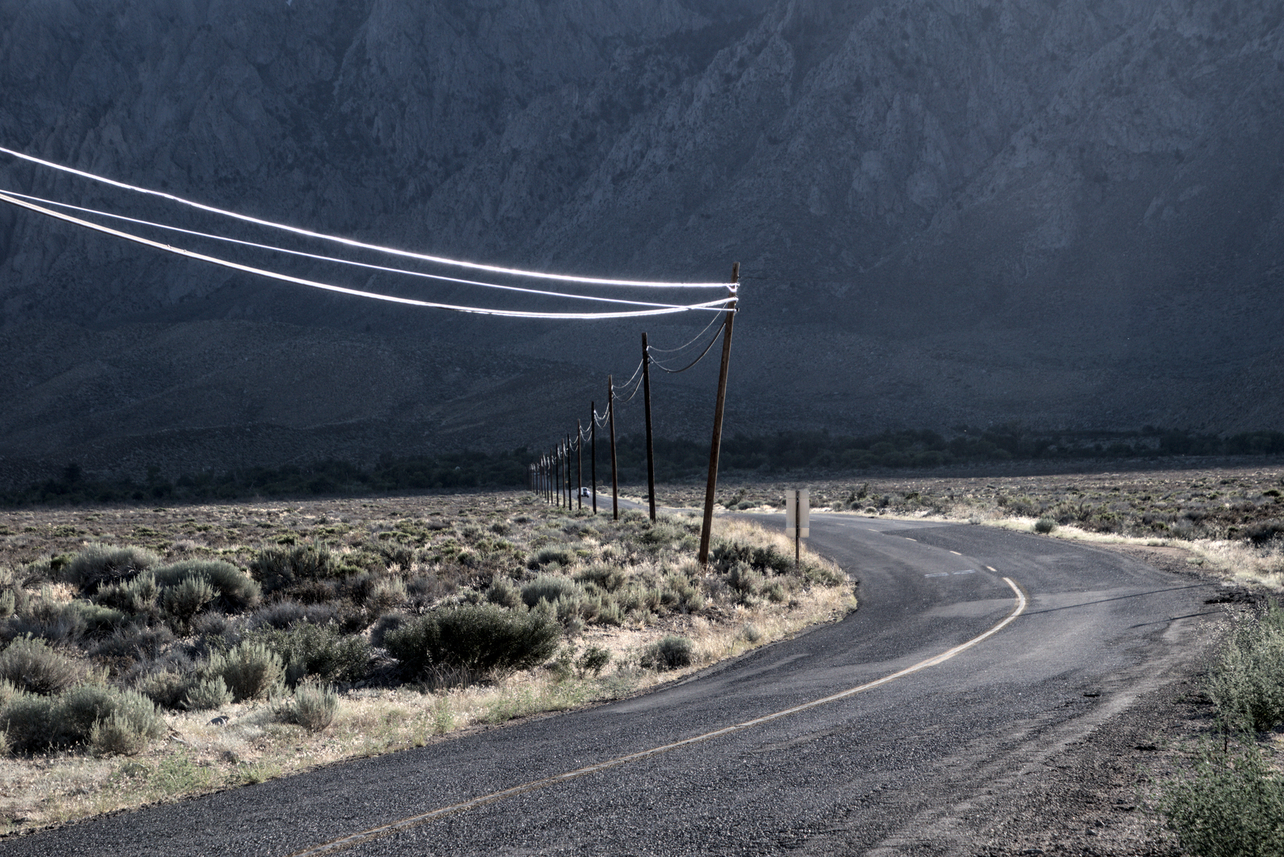 A road in the high desert banks to the left before it heads west up into a mountain canyon. The mountains in the background are in deep shadow, but the road is lit by the late afternoon sun.  The lines on the utility poles on the south side of the road reflect the sun brightly, echoing the turn in the road.