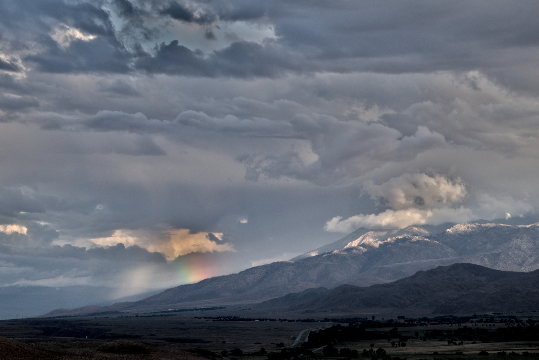 A large mountain valley is covered by storm clouds, but a ray of setting sunlight lights up a small mountain peak and creates a very short rainbow.
