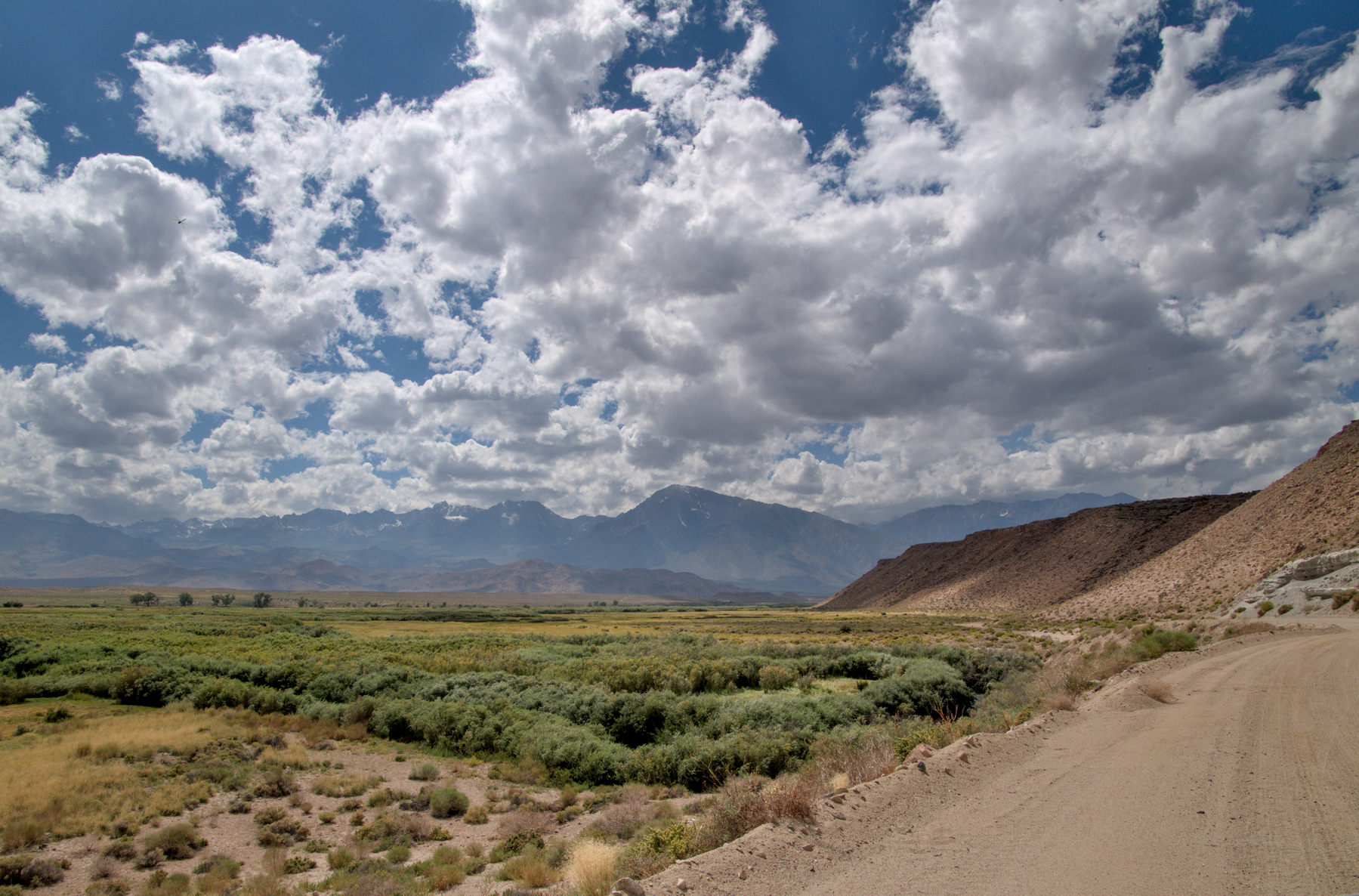 A wide green river valley is bordered on the right by a bluff of pink volcanic rock.  The Sierra Nevada is seen in the distance and the sky is full of cumulonimbus clouds.