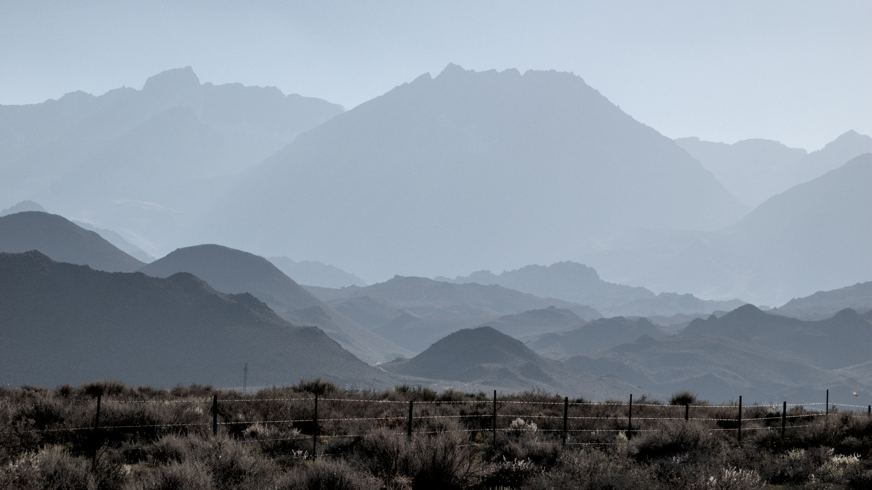 A barbed-wire fence runs along the edge of high-desert scrub, receding to rocky foothills of the Eastern Sierra.  Haze obscures the details of the hills.  The closest, lowest hills are dark grey; the highest are light grey and barely distinguishable from the faded blue sky.