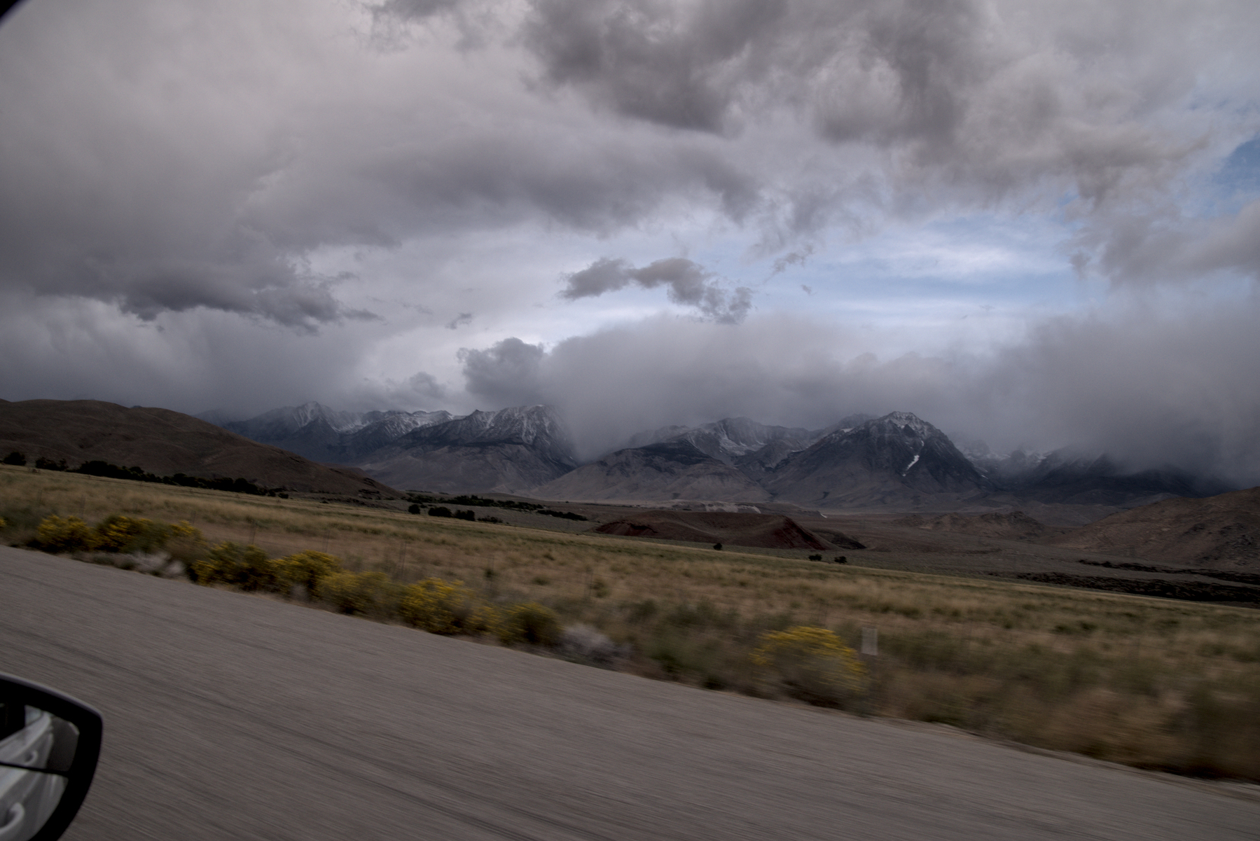 Mountains under a snowstorm, above a grassy plain, are captured from a car window.  The image is titled, a finger obscures part of the frame, the roadway is blurred.