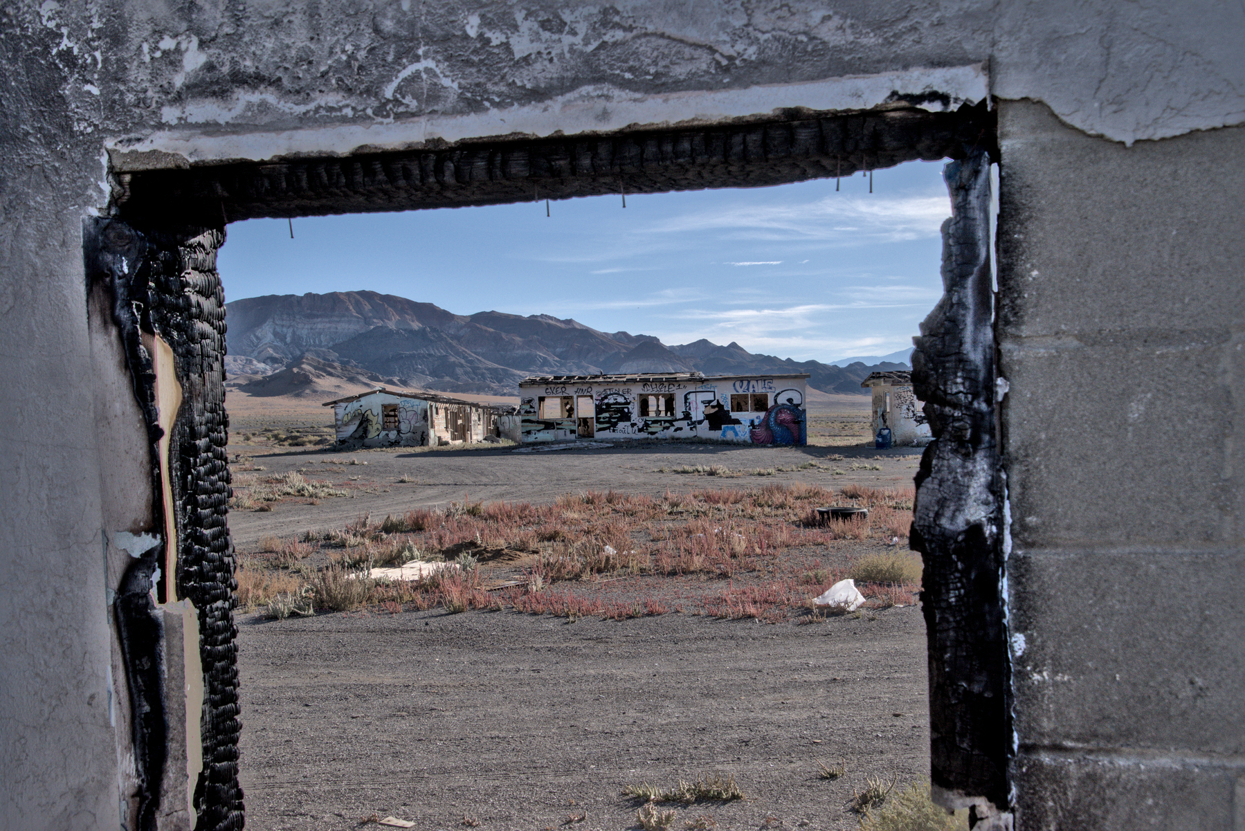 An abandoned building is seen through the door of a similarly abandoned building. The landscape is desert with mountains in the background.  Trash is spread around the ground between the two buildings.