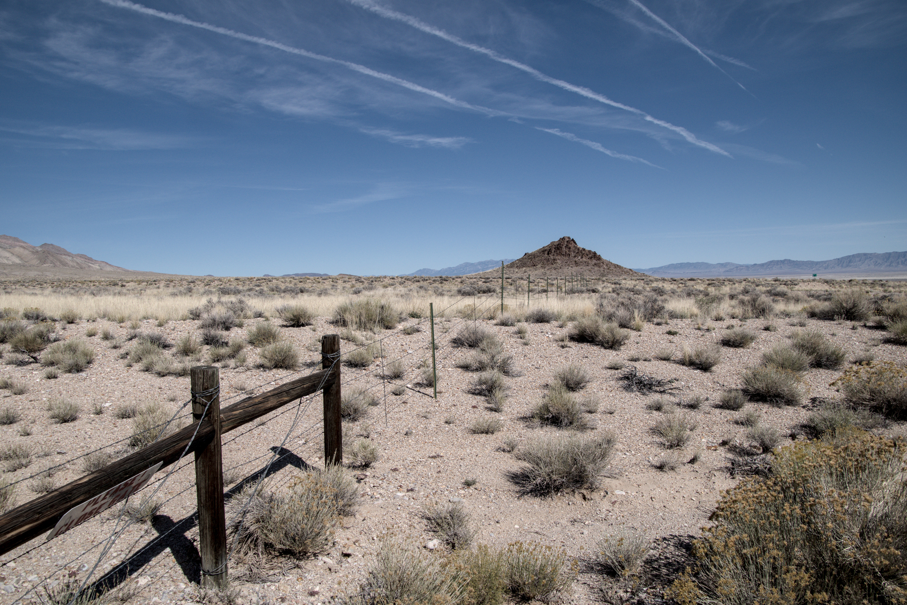 In a wide desert valley, a wooden and barbed-wire fence runs towards a cone-shaped hill in the valley. Airplane contrails in the sky parallel the fence.