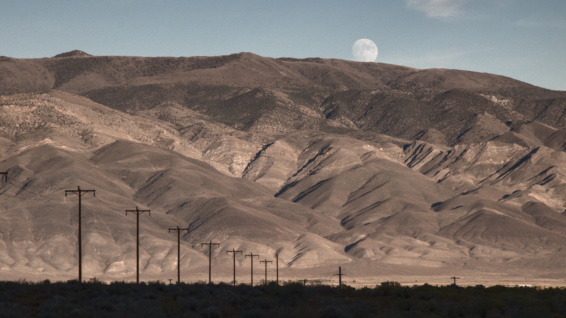 Power poles, near on the left, recede towards a high mountain range, which is eroded and barren of greenery, so that it seems soft and brown.  A nearly full moon has risen just above the mountain ridge-line.