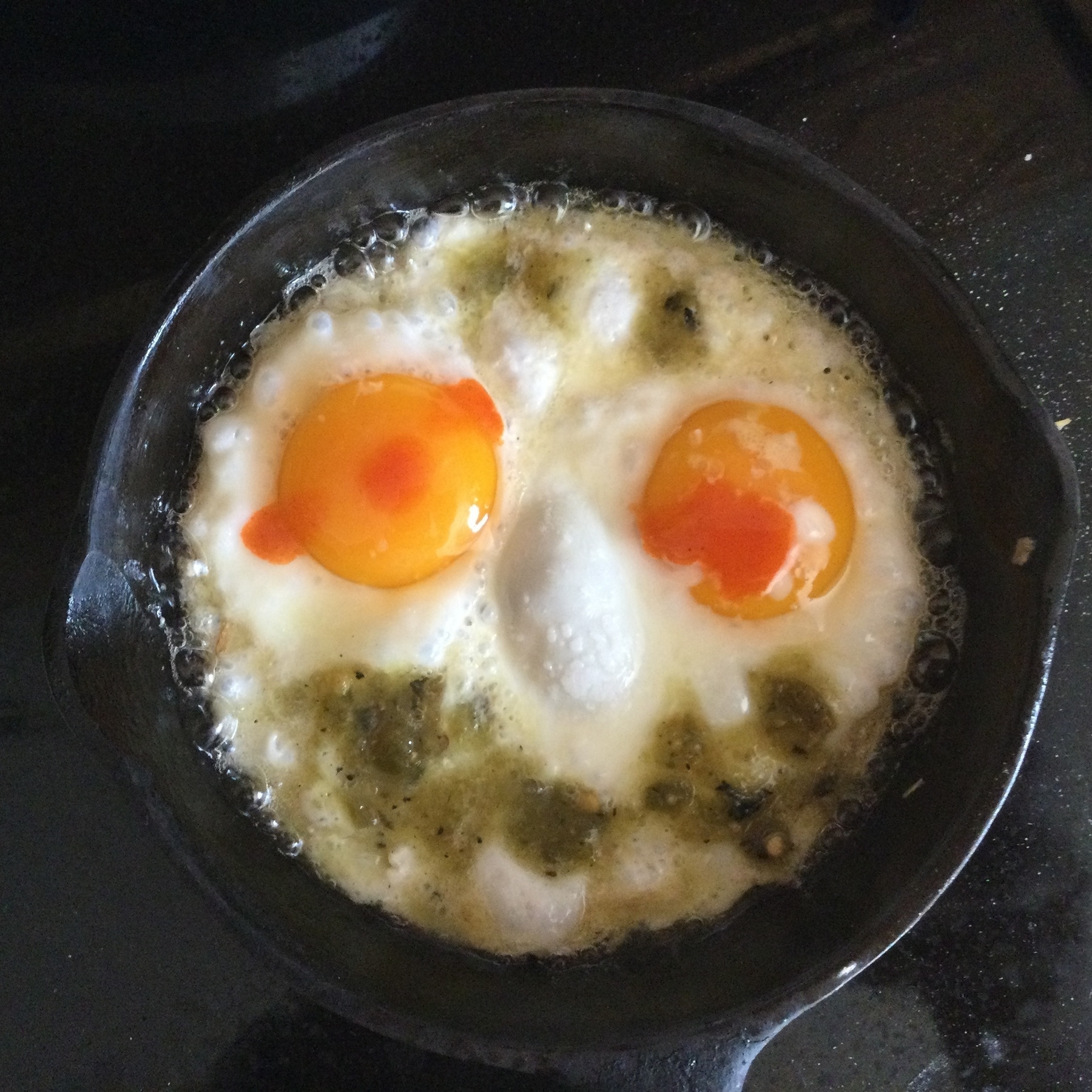 Two fried eggs in a cast iron pan, the yolks look like eyes, a bubble of egg white looks like a nose, an arc of grfeen salsa forms the mouth, and each eye is dotted with red hot sauce.