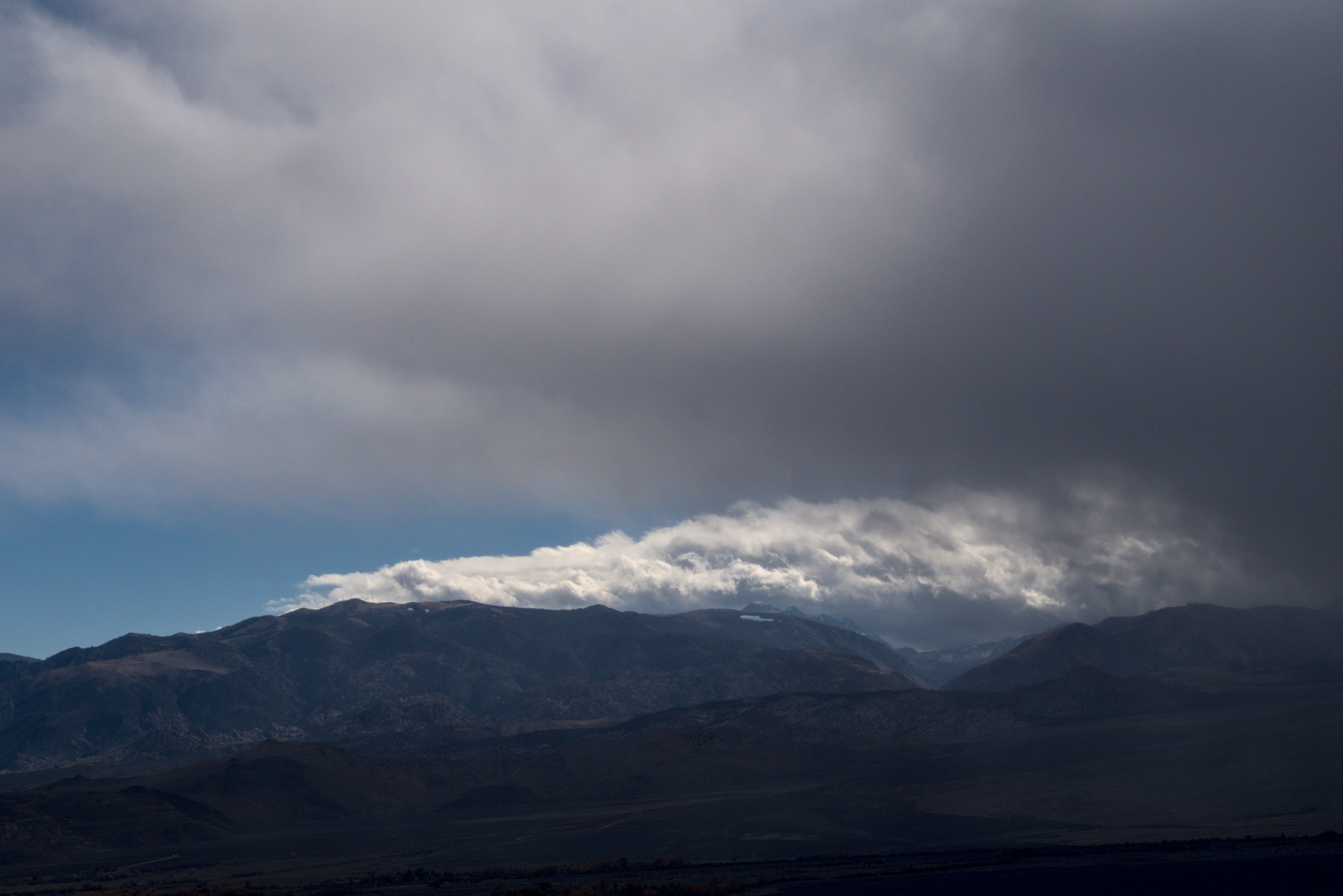 A narrow band of puffy white clouds and blue sky can be seen through a gap over some mountains and under storm clouds.