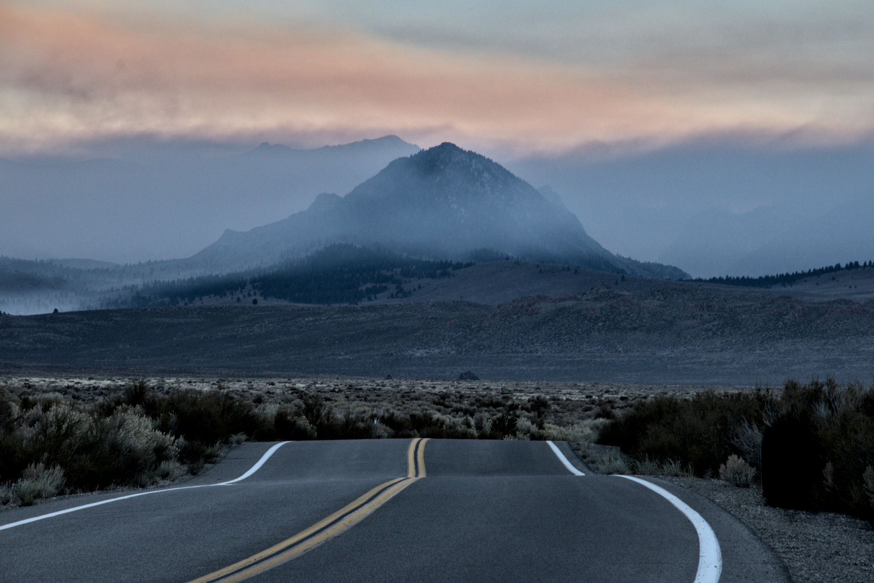 A rolling highway in the foreground opens up to a large valley surrounded by mountains, with a volcanic cinder cone in the middle.  Smoke drifts in from the left, partially obscuring the cinder cone.