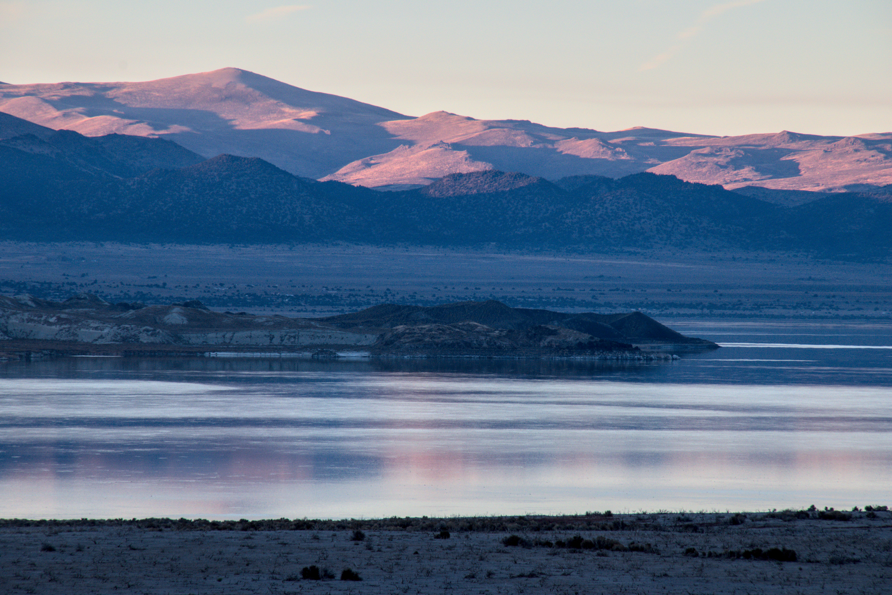 A large lake is calm as the sun sets, lit with pinks and blues from the surrounding hills.