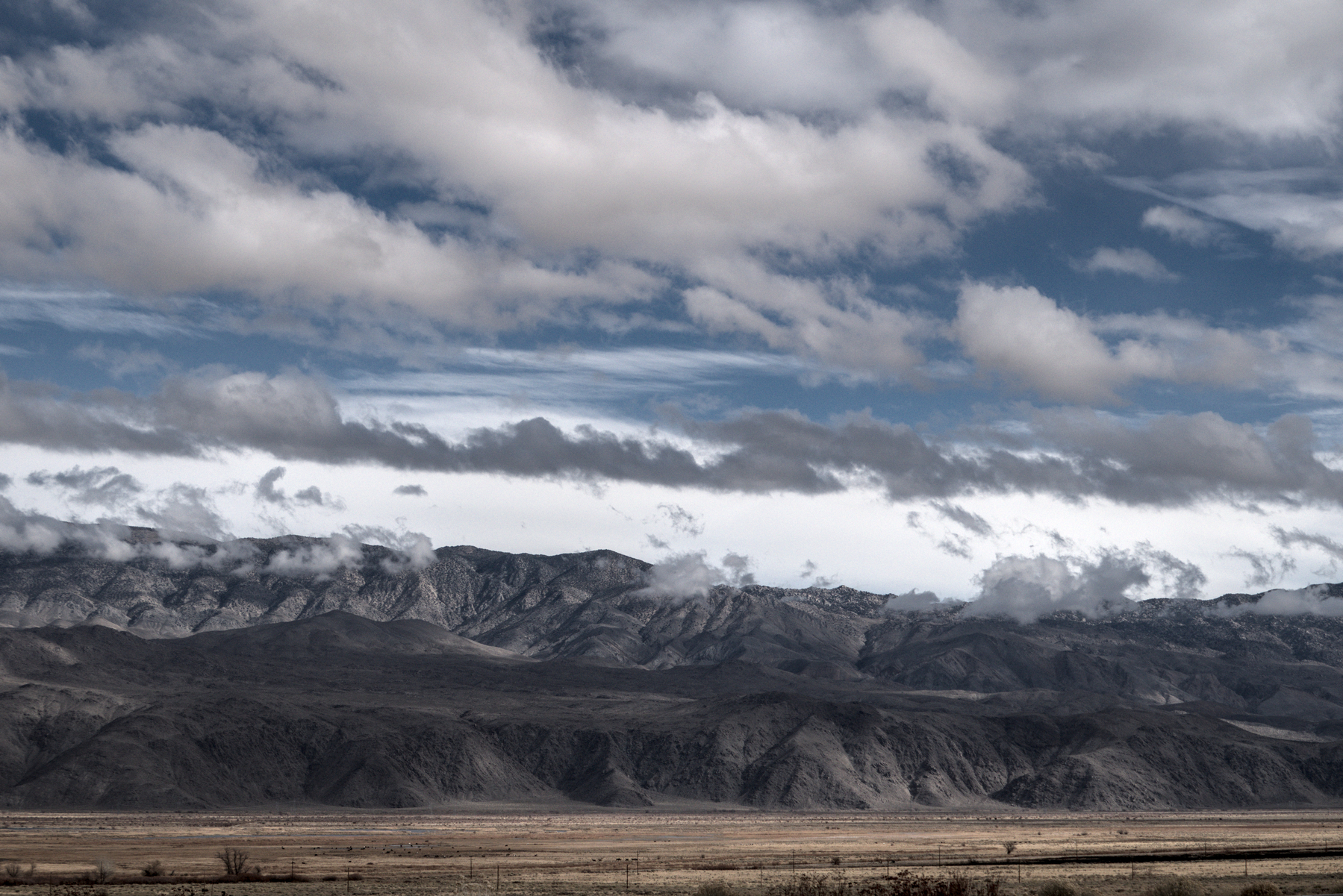Grey-brown weathered mountains loom over a grassy valley, with clouds on the mountain peaks and above casting shadows.