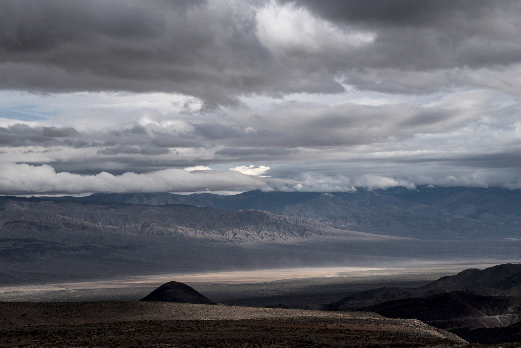 Looking down from a high point, a large, barren valley runs left and rihght. 
 Light hits the bottom of the valley, while the foreground is mostly dark.  Patches of light hit the rugged, eroded mountains on the far side of the valley. Clouds fill the sky, but not uniformly, there are many different types, and patches of blue show through.