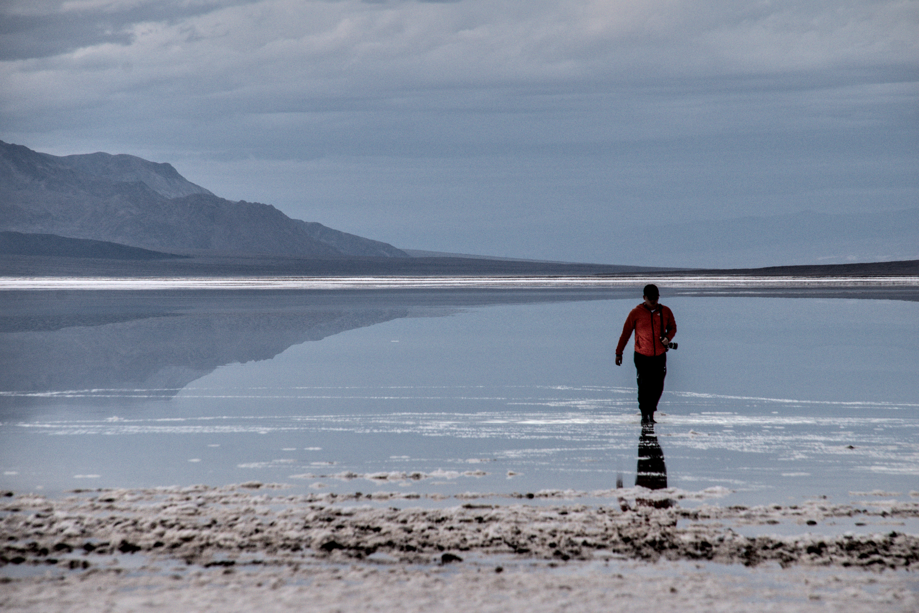 A man appears to walk on the surface of a large lake. The surface is smooth, reflecting the mountains in the distance.