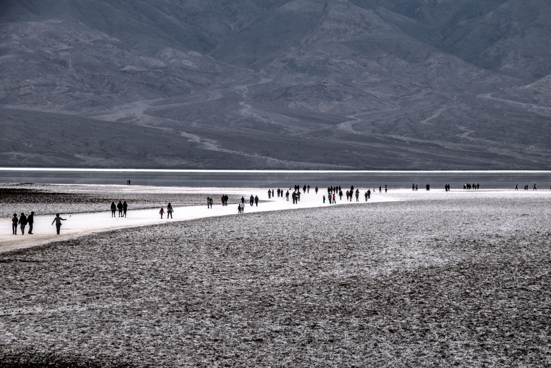 A long, but sparse, line of people walk along a white alkali path to the edge of a shallow lake. Dusky mountains rise from the far side.  Everything is brown or white.