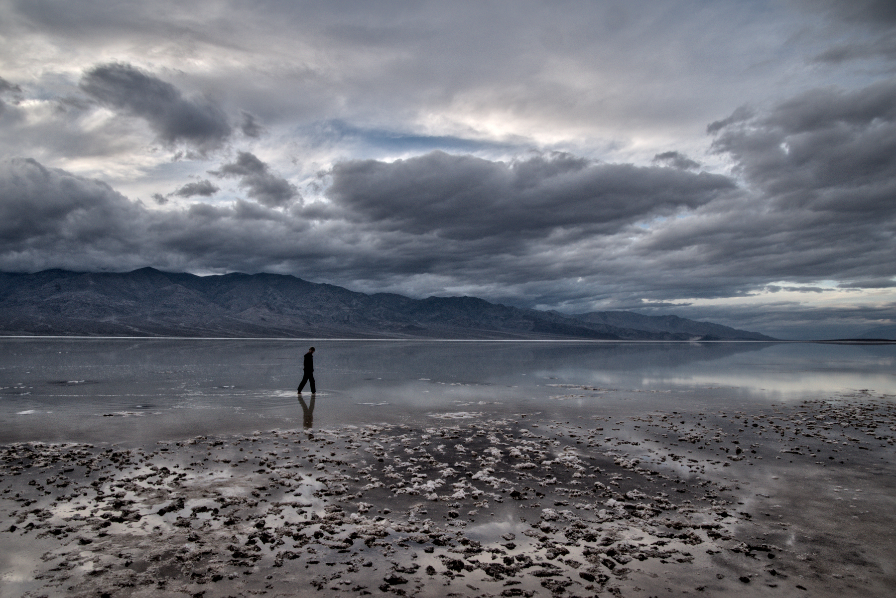 A man walks through the shallows of a shallow, salty lake.  There shores are white salt, dusky mountains rise from the far shore.
