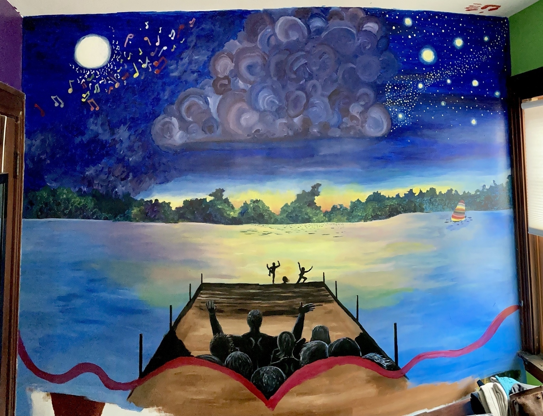 A wall is painted in bright colors depicting a sunset at a lake. Children jump off a dock while adults watch on.  A sailboat on the lake has a many-colored sail.  Clouds, stars, and the moon fill the twilight sky above.