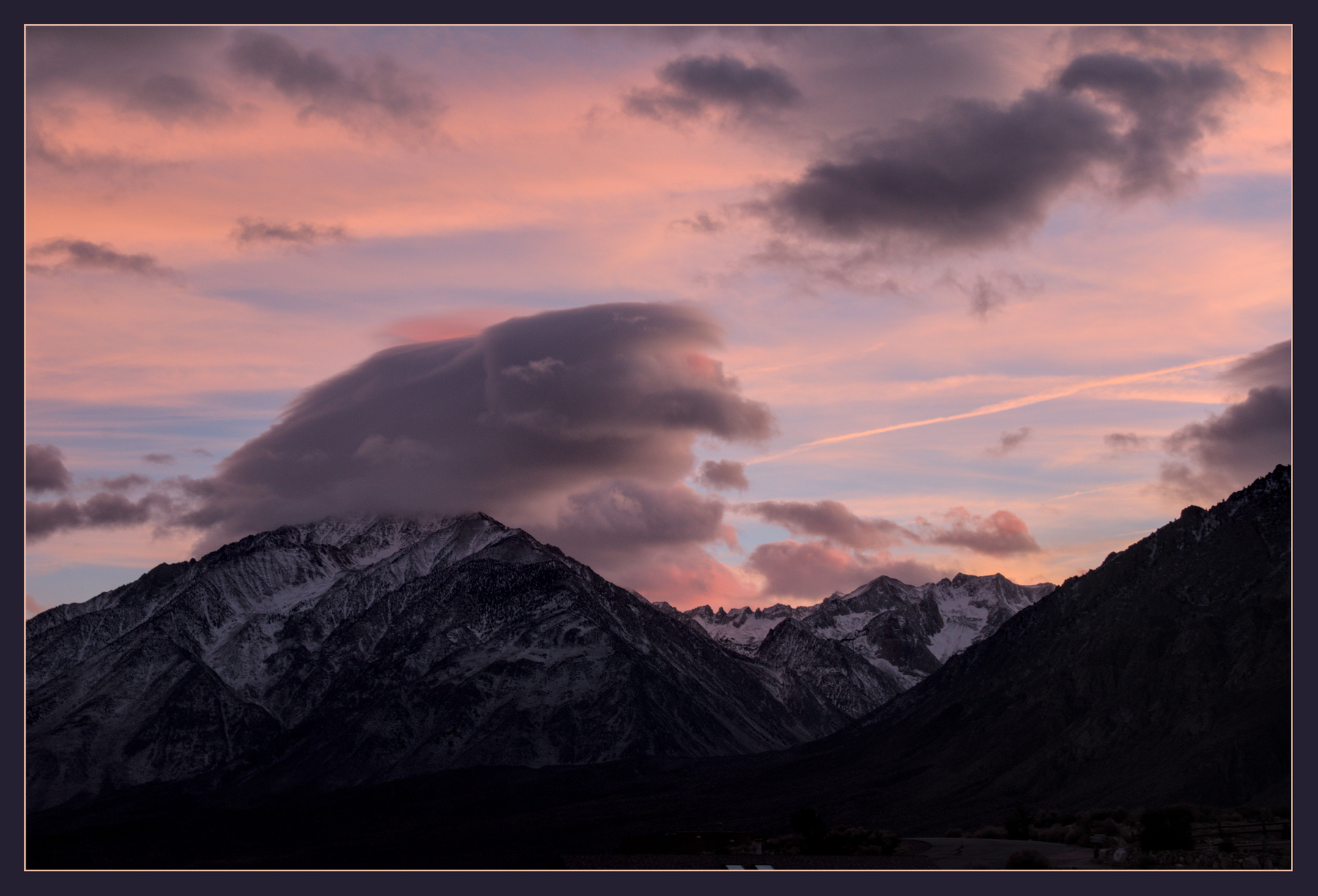 A triangular mountain with  partial snow cover has a large puffball of a cloud sitting on its peak.  The sky behind has high wispy clouds, turned pink by the setting sun, while all the lower clouds are grey.