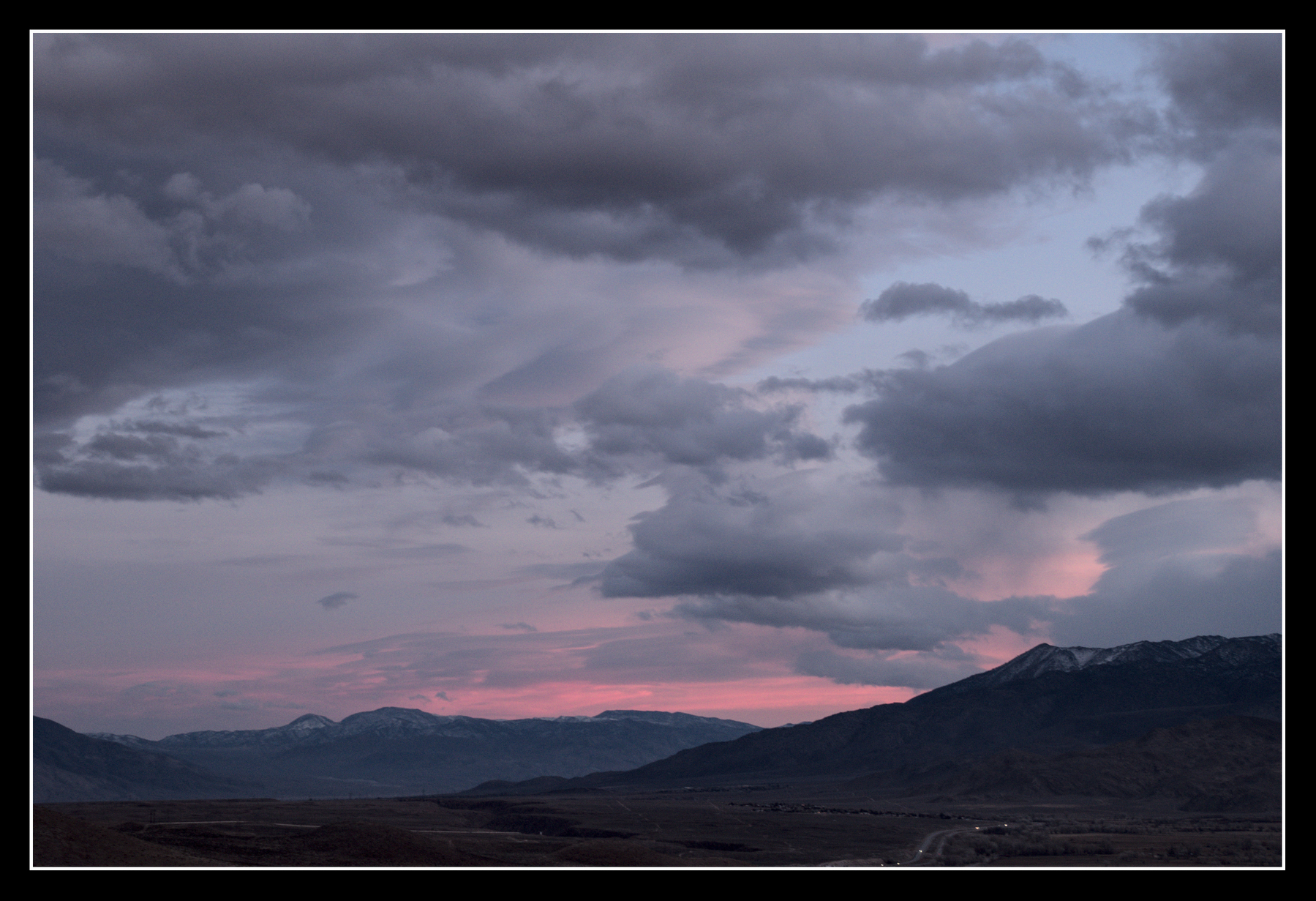 A broad valley surrounded by mountains has many varied clouds hovering over it.  The sunset has lit some pink, some blue, some grey, and seemingly every shade between.