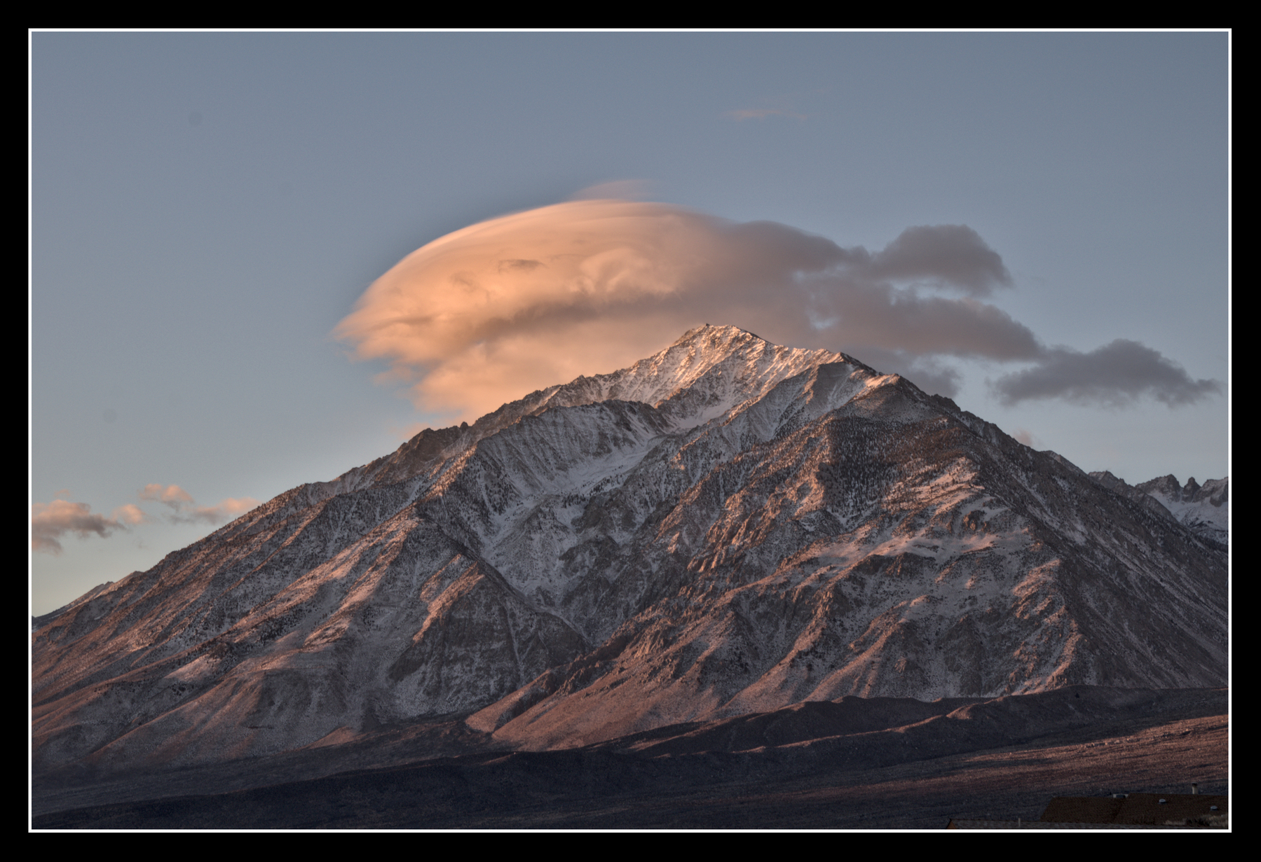 A triangular, snow-capped mountain is lit by sunrise.  A lenticular cloud sits on top of it.