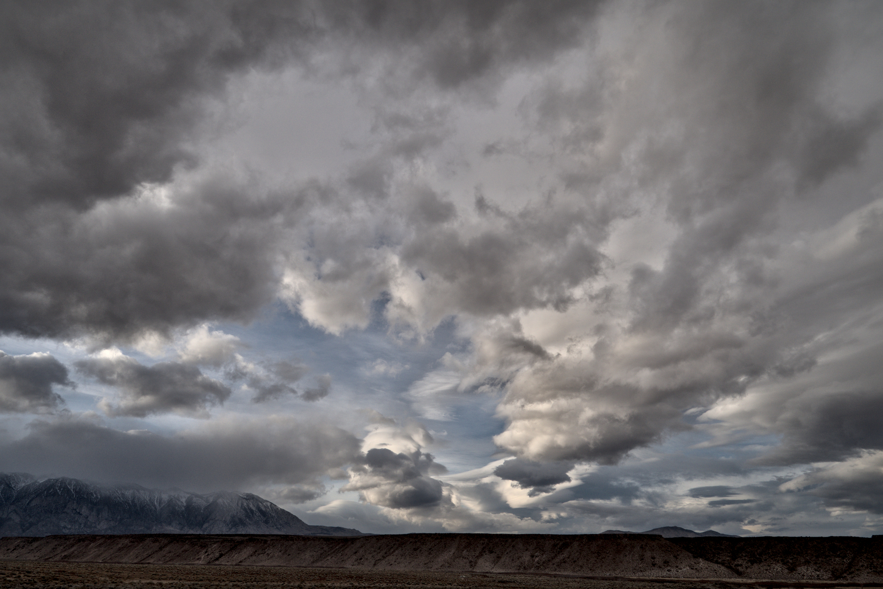 A wide shot of the sky above mountains has many different types of clouds, some white, some grey, with patches of blue sky between.