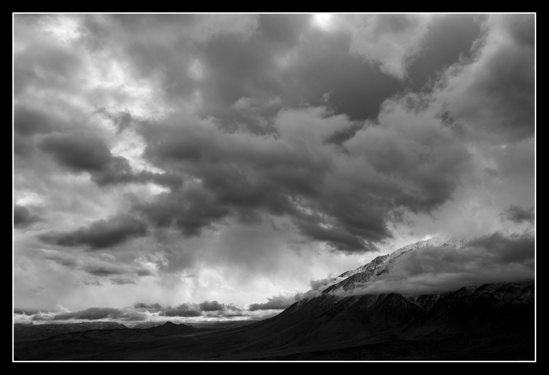 A long mountain ridge lies beneath a sky full of clouds varying in form and mostly dark shades of grey.