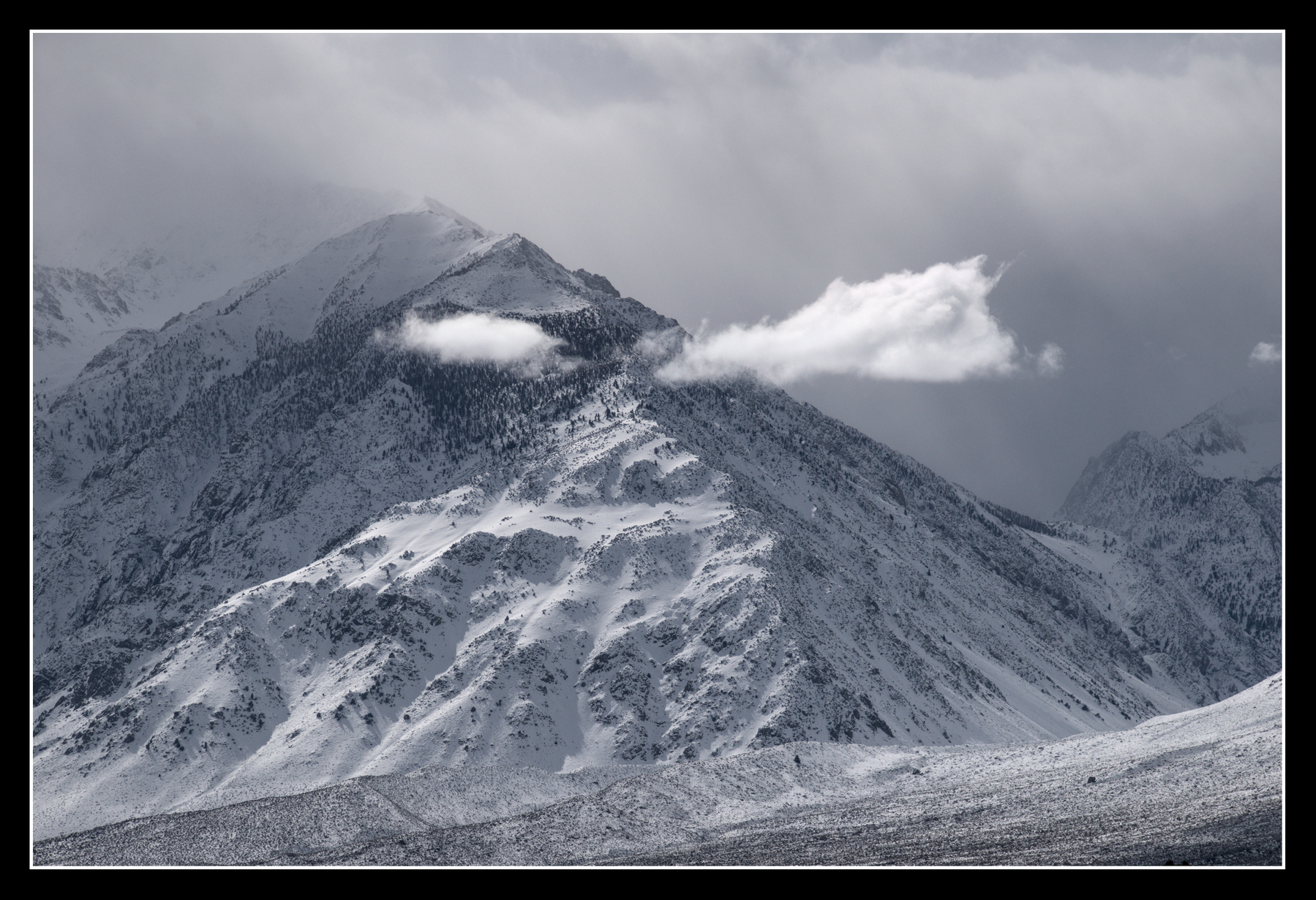 A snow-covered mountain has a sharp ridge line running from summit to ground. Two small clouds on either side of the ridge line make it look like a face, with the clouds as eyebrows.