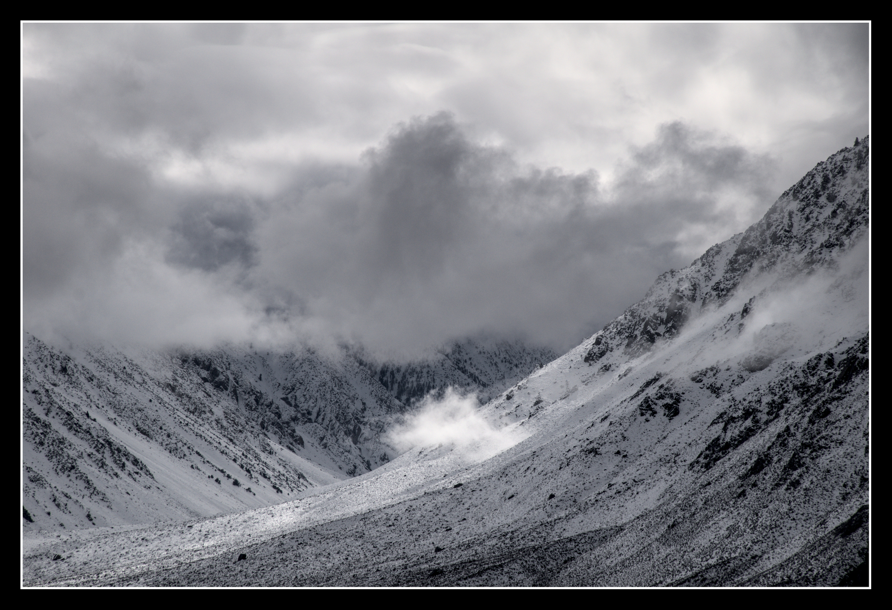 A deep. sharp mountain canyon is covered with snow. Clouds cover the tops of the mountains, but one little cloud rests on the canyon wall, apart from the rest, and lit by a ray of sunlight.