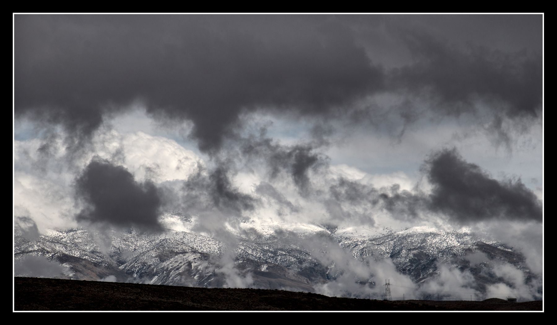 A mountain range runs across the bottom of the image, partially covered in snow.  Fragmented, dark clouds hover near the mountains, while puffy white ones rise in the distance.