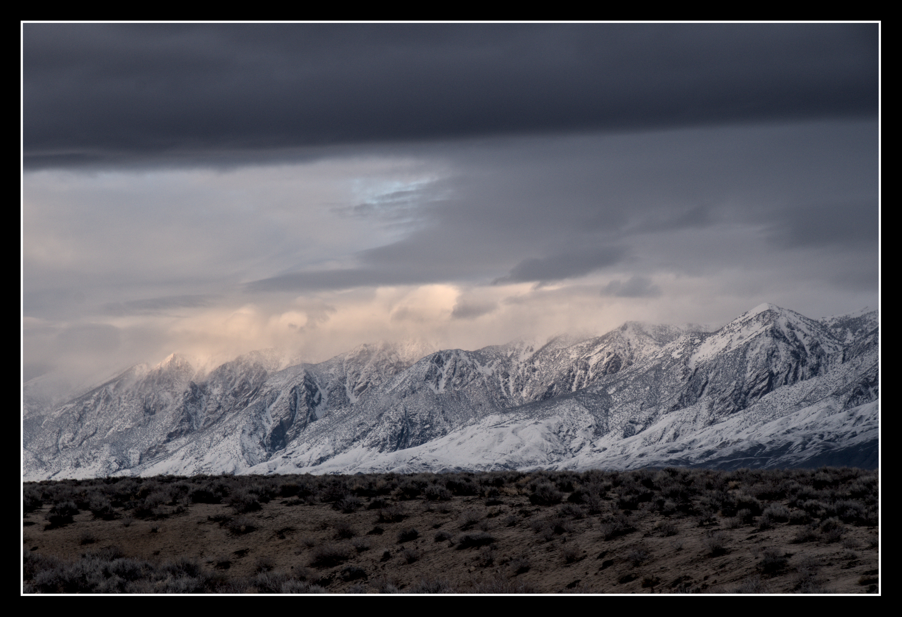 A wide, snow-covered mountain ridge is sandwiched between a dark layer of clouds above and high desert scrub below.