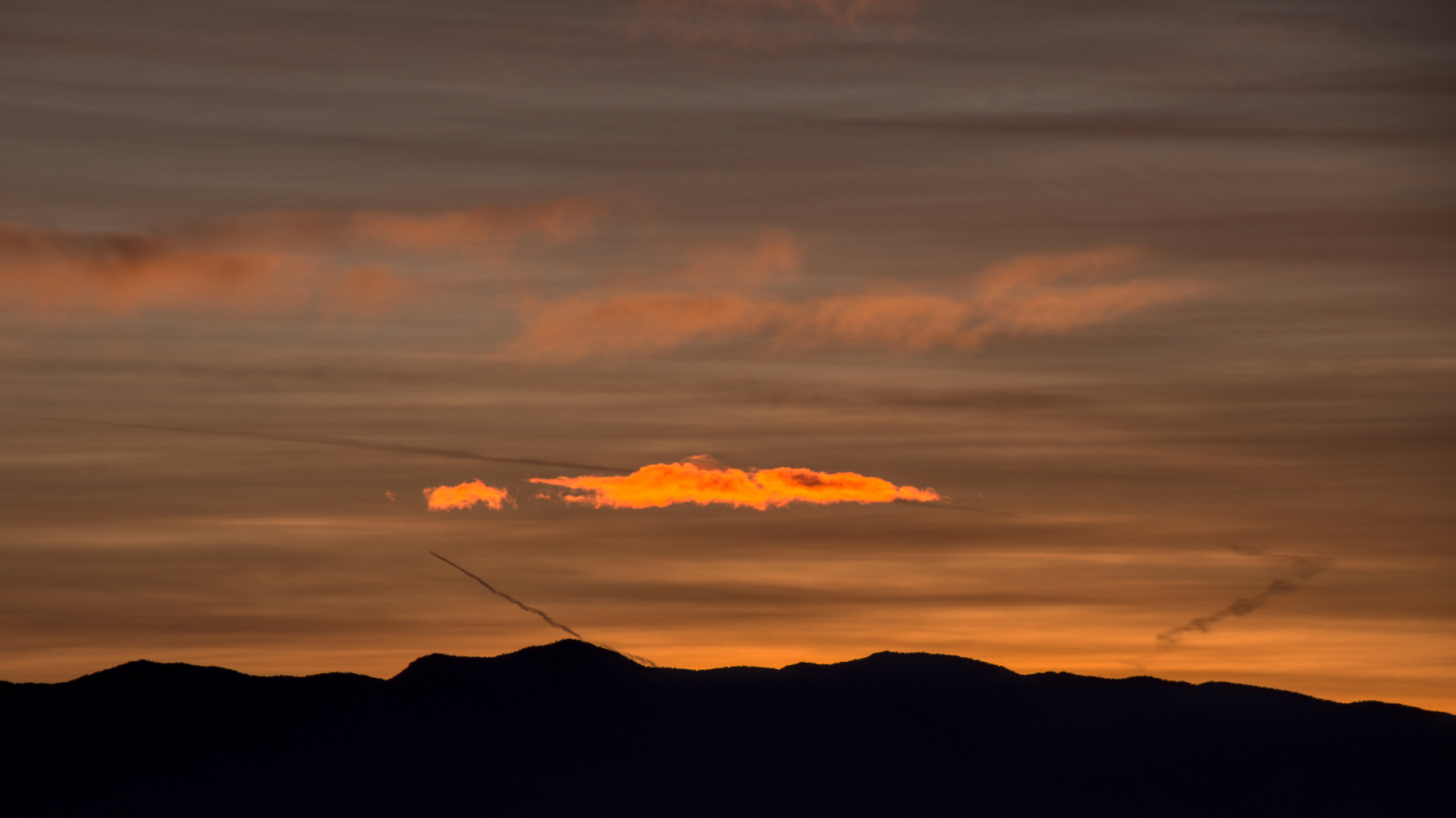 A mountain range blocks the sunrise, but clouds above it are brightly lit from behind.