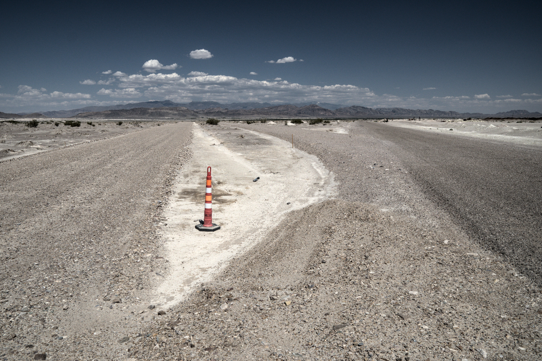 A skinny traffic cone marks a bit of rough white sand between two gravel roads through the desert.