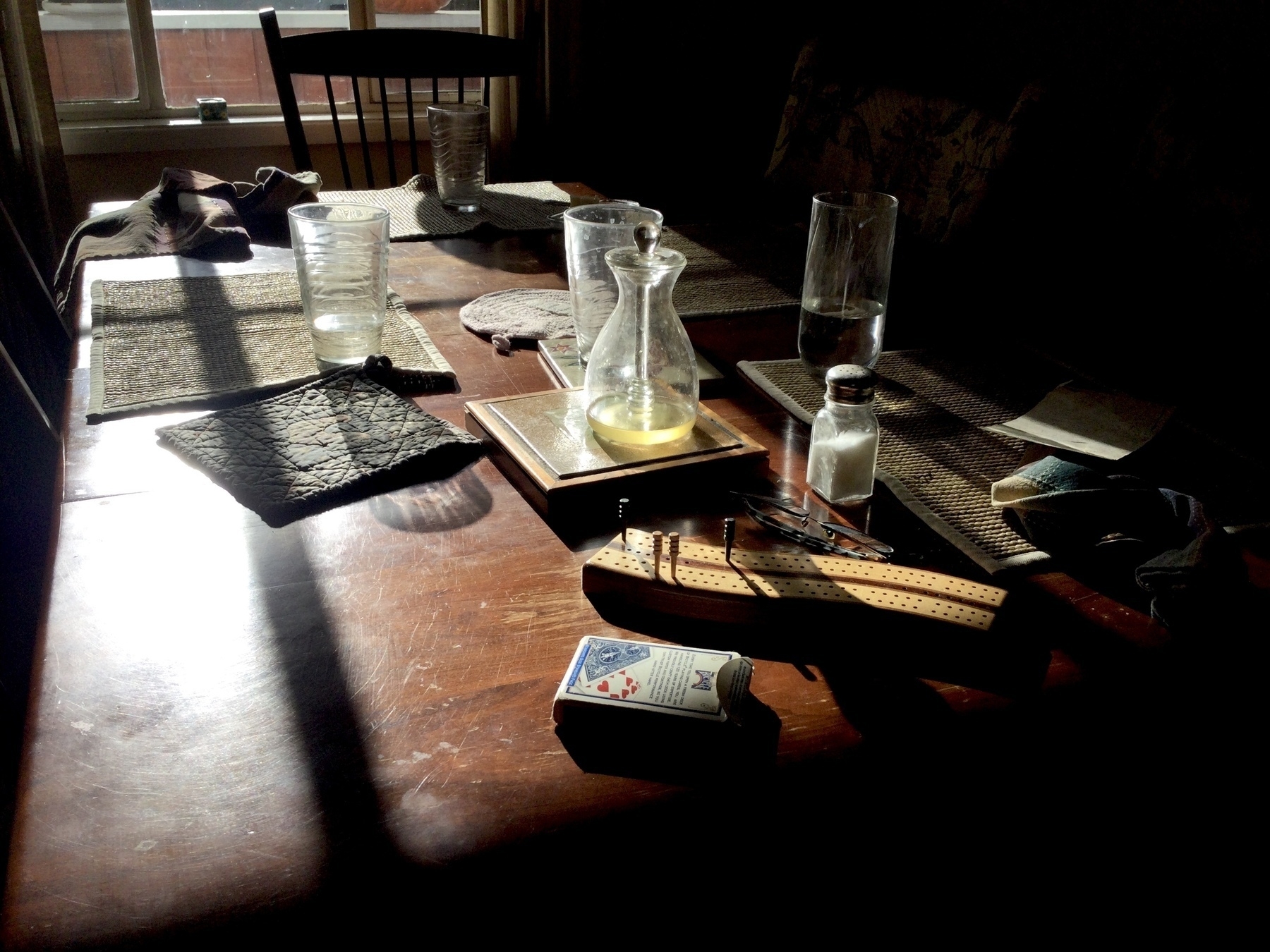 Morning light streams through onto a dining table with water glasses, place mats, napkins, salt shaker, a glass honey pot, a deck of cards, and a cribbage board. 