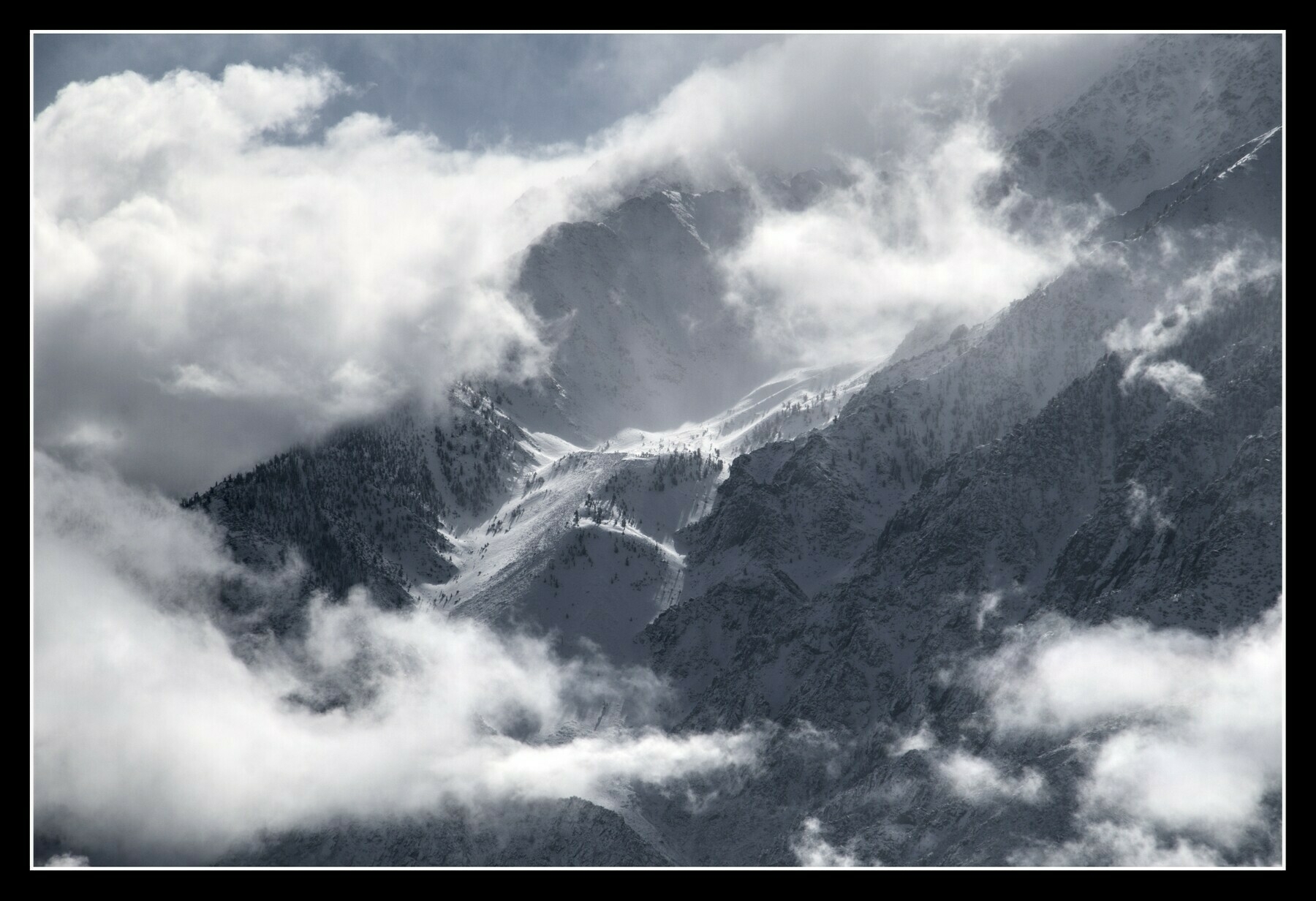 A snow filled canyon runs from the top of a jagged mountain, partially obscured by clouds.