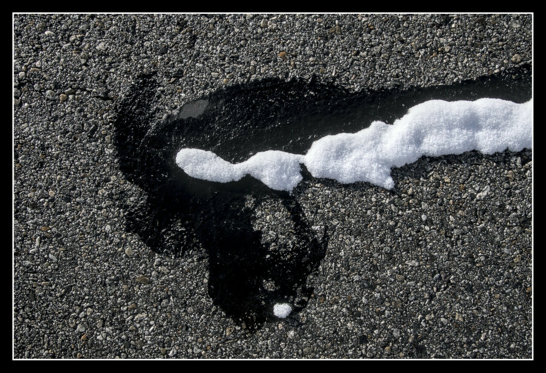 Looking down at a patch of tar on a road, snow has melted at the edges of the tar, still covering the center.