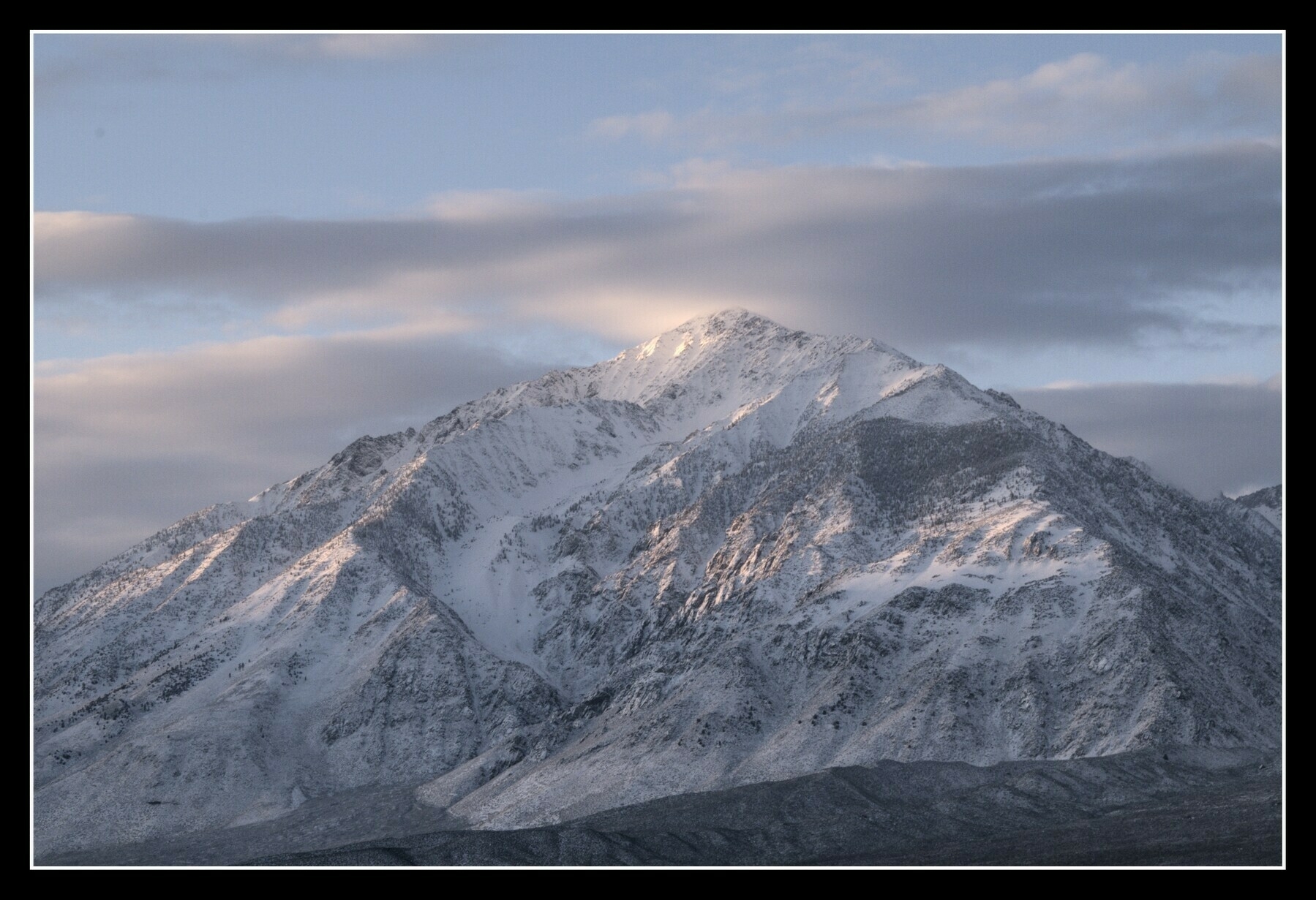 A triangular, jagged mountain, covered with snow, is lit pink with light from the rising sun.