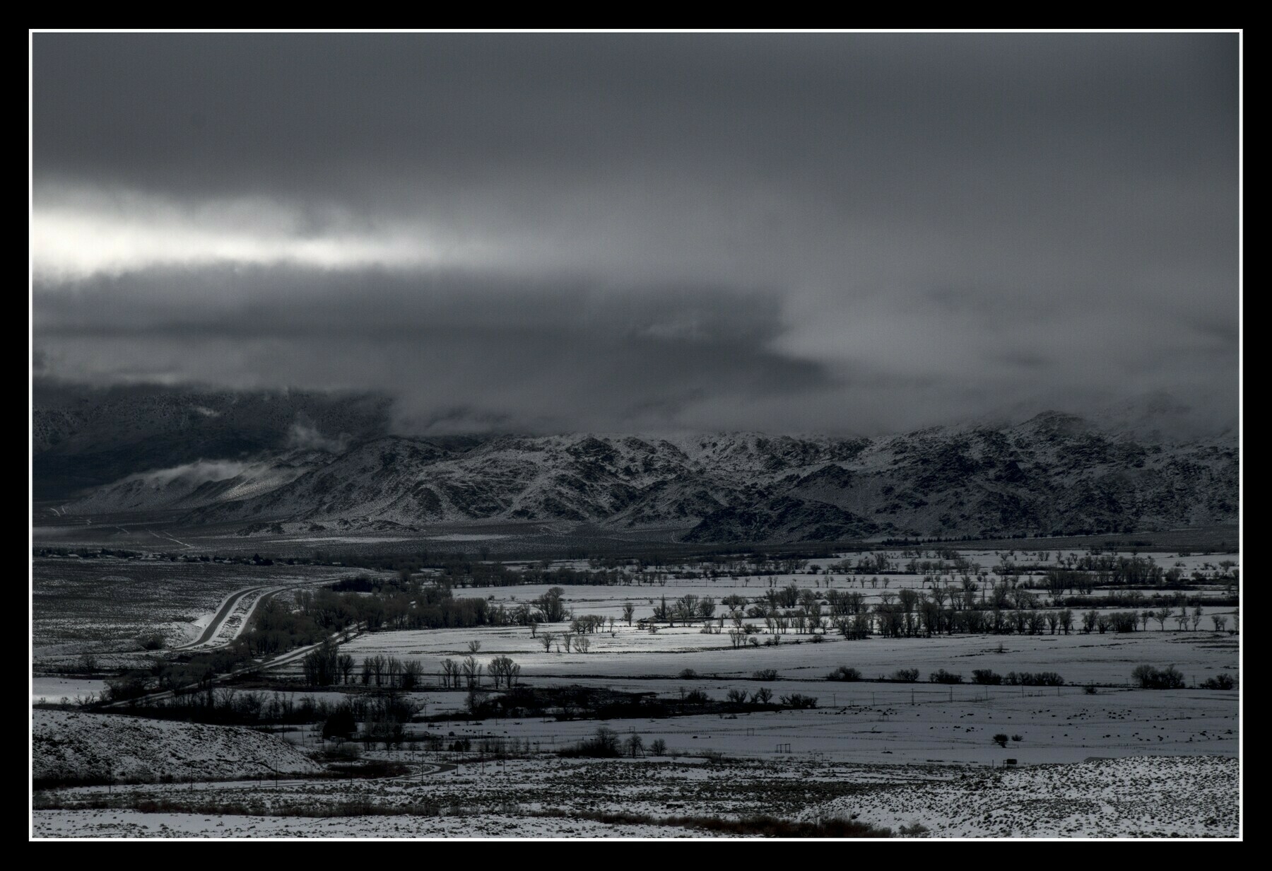 A valley floor has fresh snow, lit through a hole in the overhanging clouds.