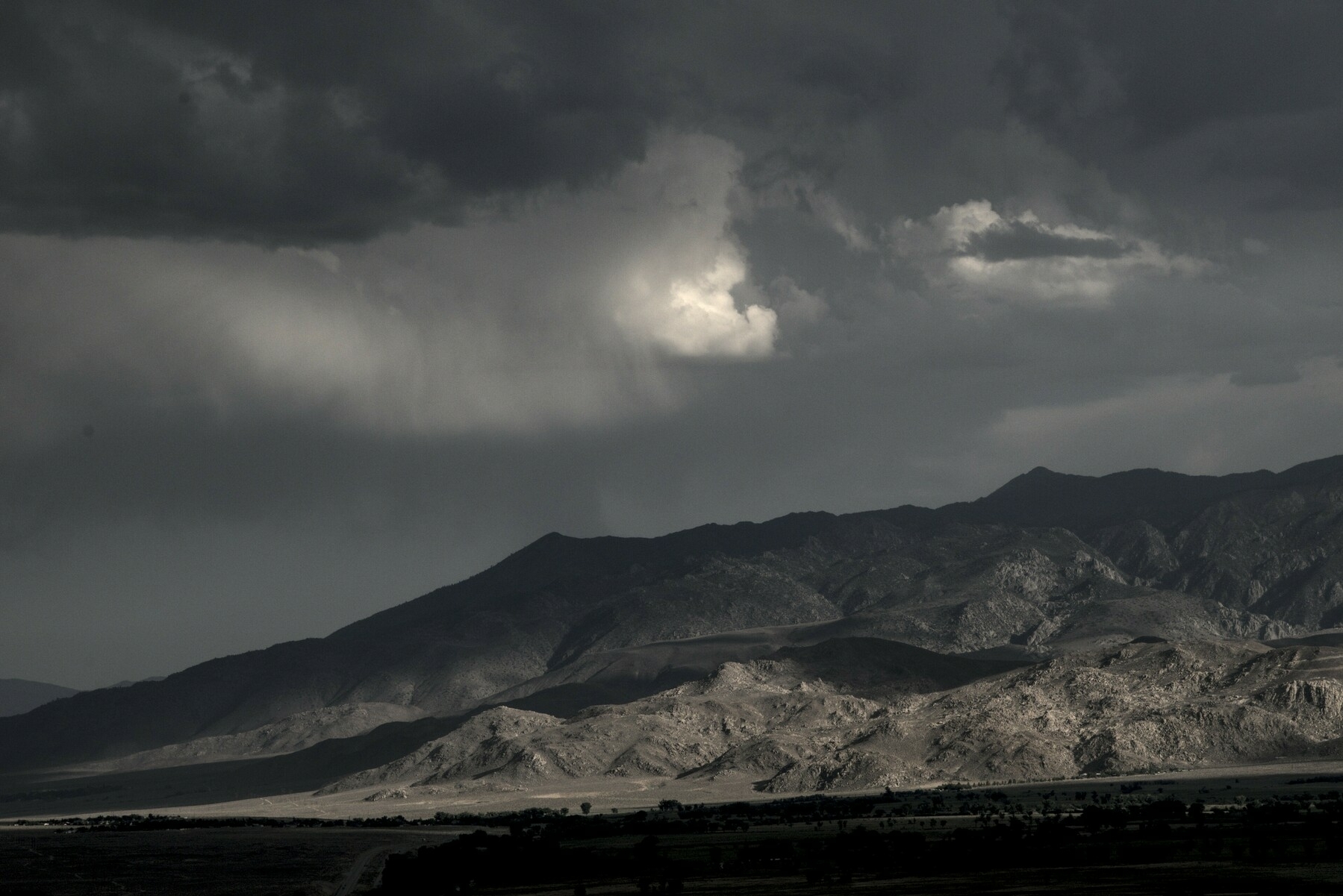 Low foothills stand out in a beam of slanting sunlight below dark clouds.