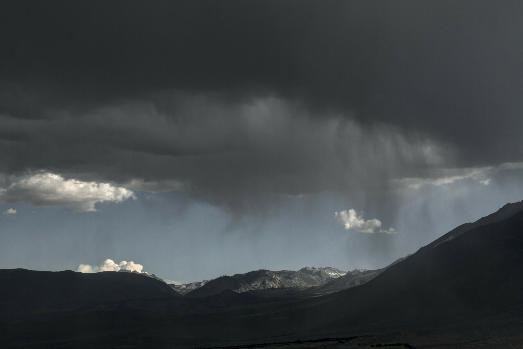 Dark storm clouds float over dark mountains, with wispy grey falling rain coming down through a gap of blue skies and puffy white clouds.