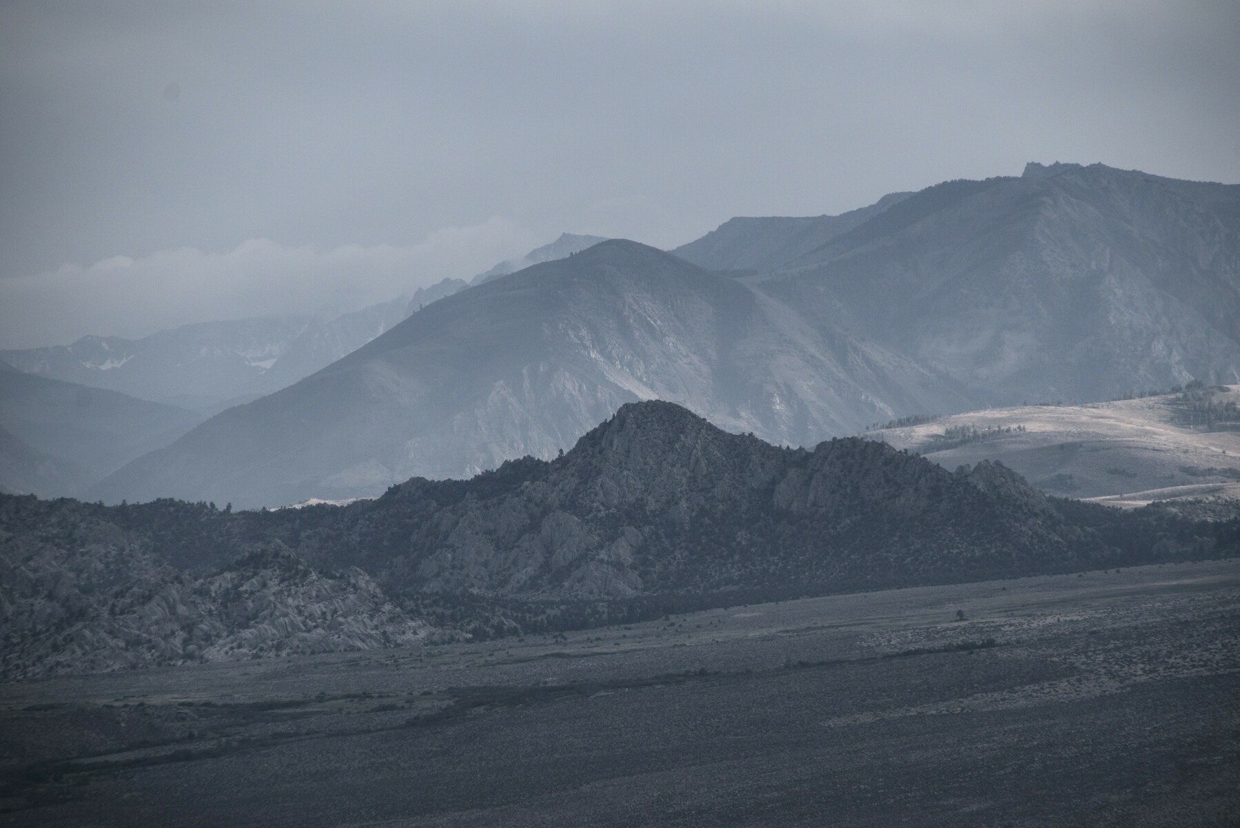 Low mountain foothills are dark n shadow, surrounded by lighter valley floor and mountains.