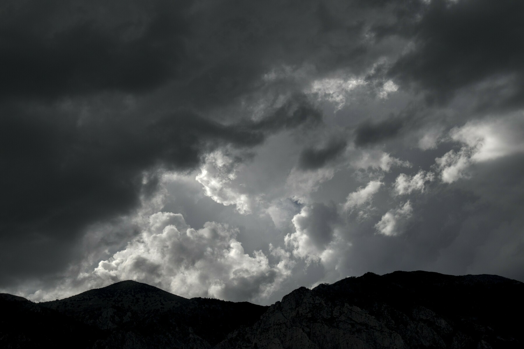 Dark clouds over mountains have a gap of a tiny bit of blue sky and lighter clouds.
