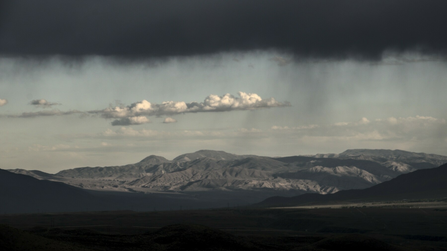 Rain falls from dark clouds over a high desert valley, while the view under the clouds shows clear skies in the distance.