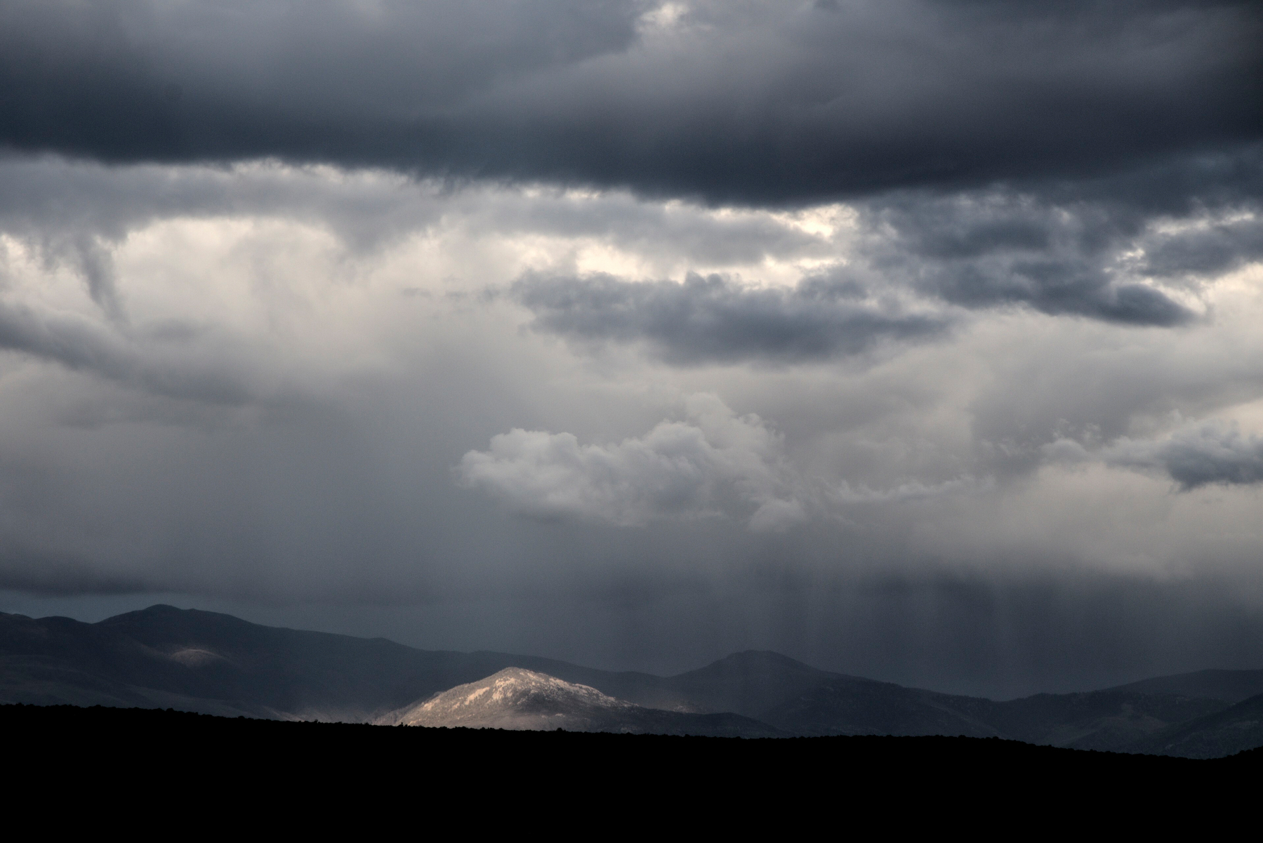 A small hill in the high desert is lit by a beam of sunlight shining through a hole in a stormy sky.