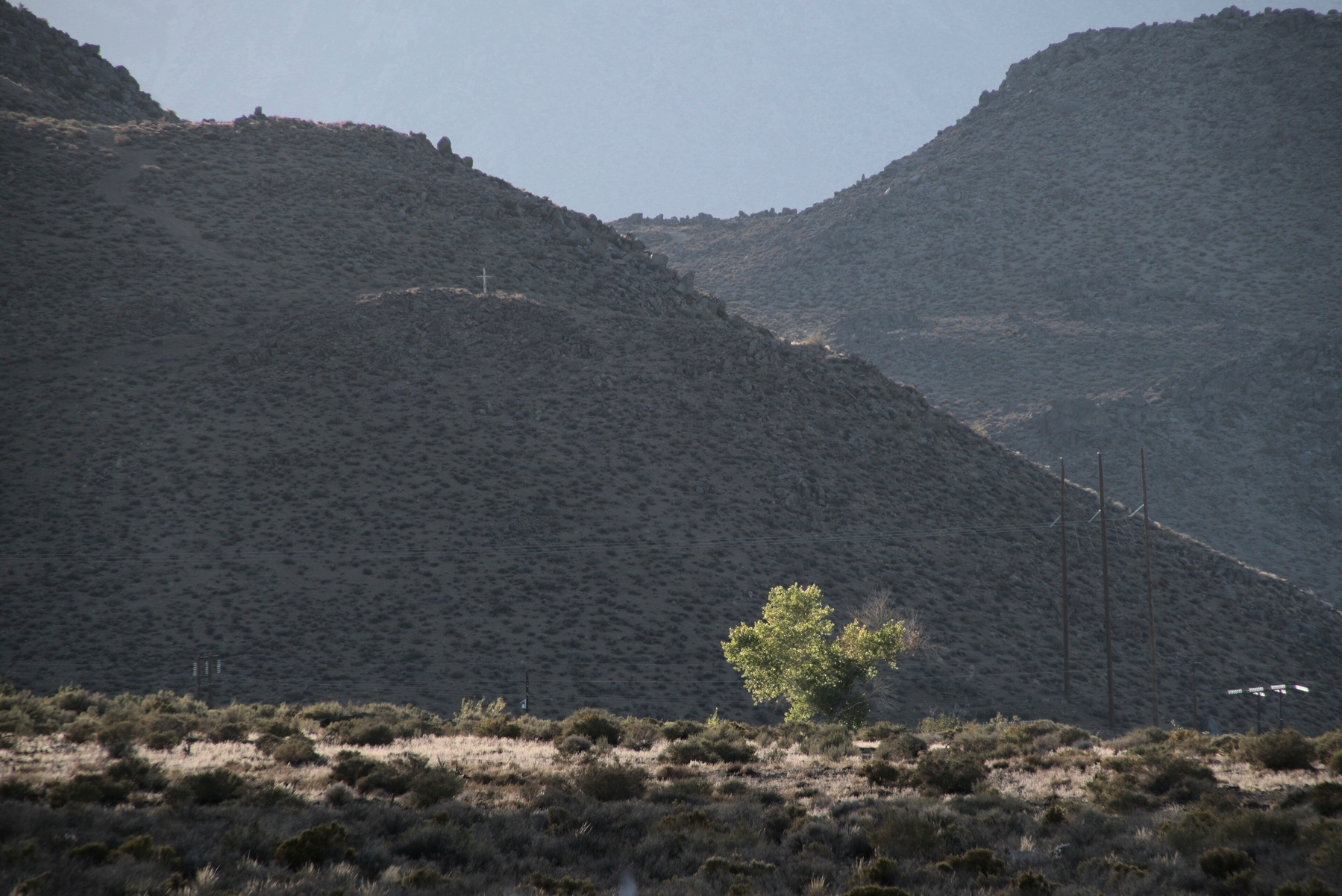 A dark mountain landscape is offset by a beam of light piercing dark clouds and lighting a tree standing by itself.