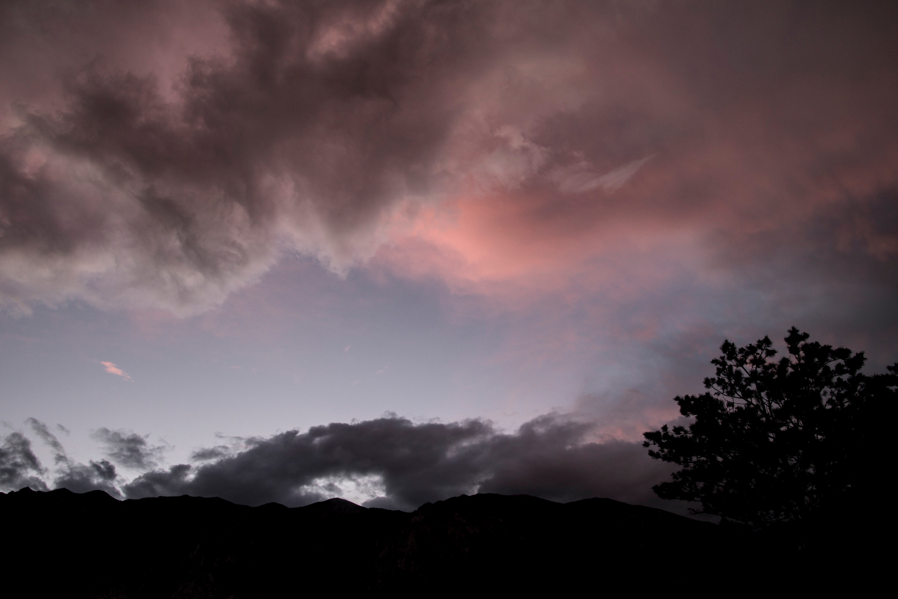 Puffy clouds over a mountain are lit pink and grey by the sunset, with gaps of blue sky.