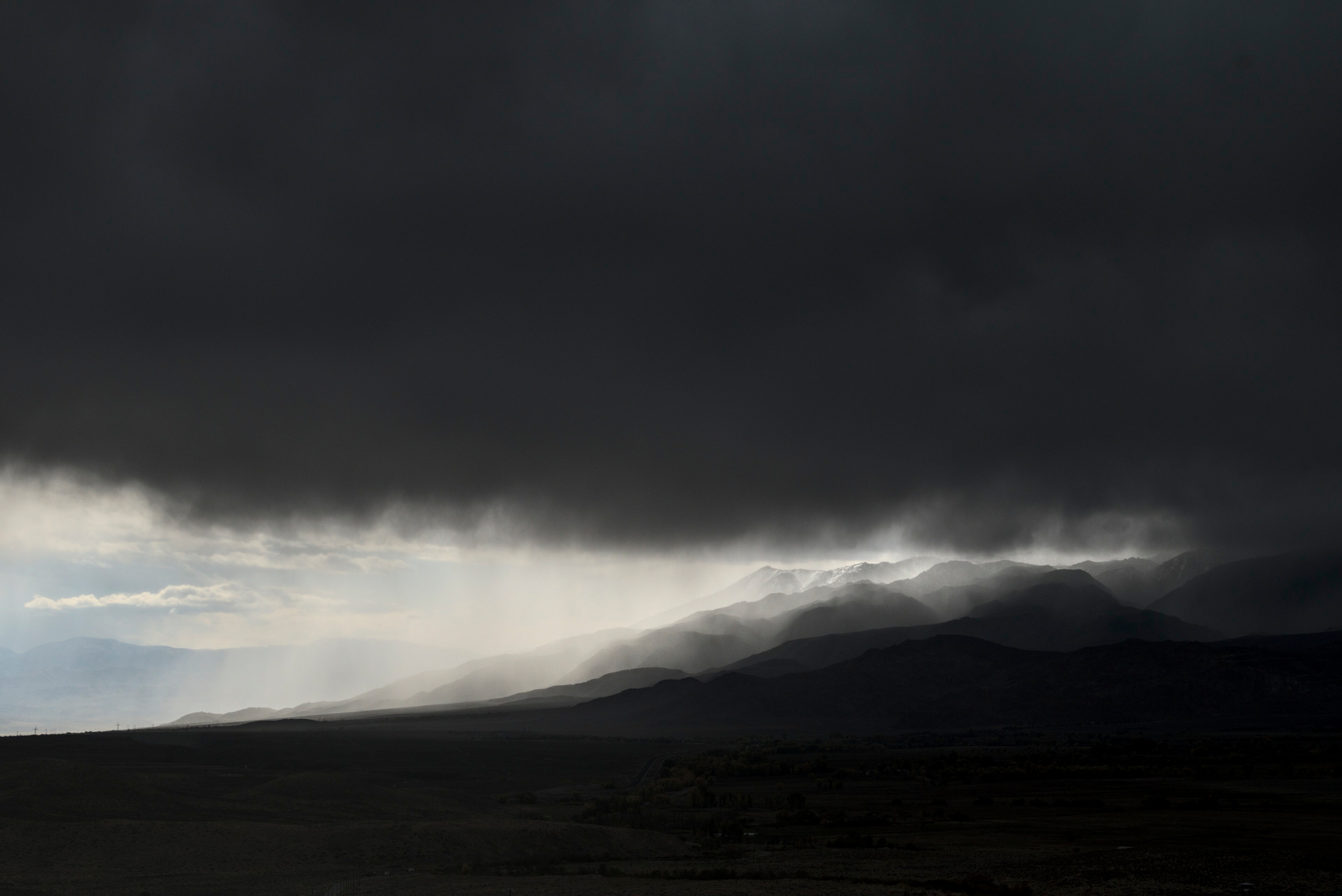 A dark cloud bank fills the sky with just a wedge of light under the cloud where the mountain foothills meet the desert.