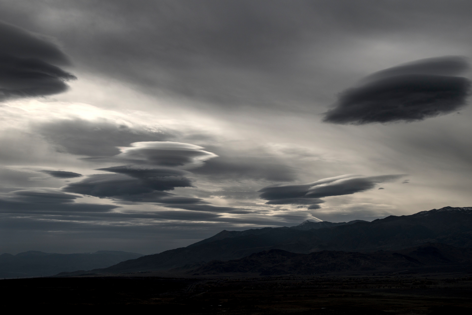A mountain valley is covered by a grey sky with lenticular clouds dotted here and there.