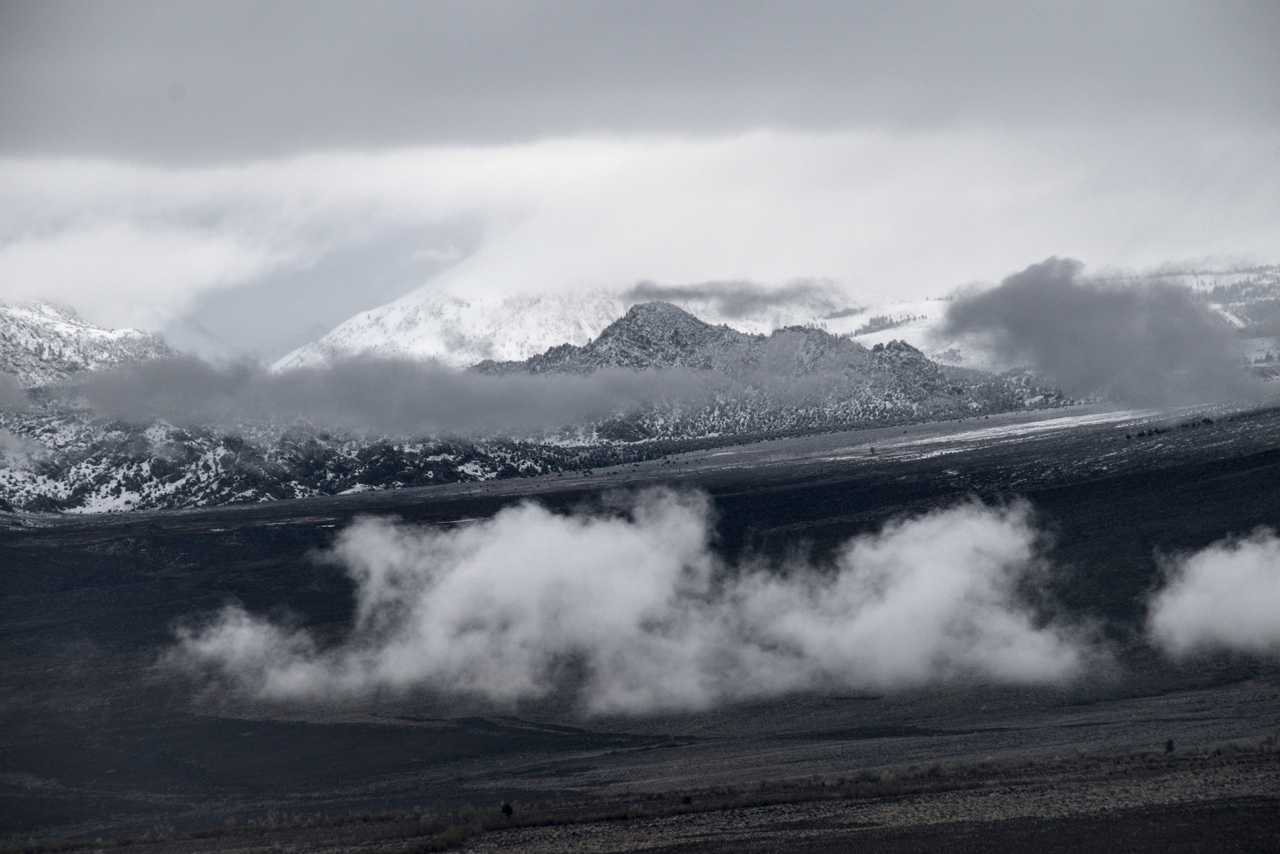 A chain of mountain foothills runs below mountains and clouds, above a lower valley.