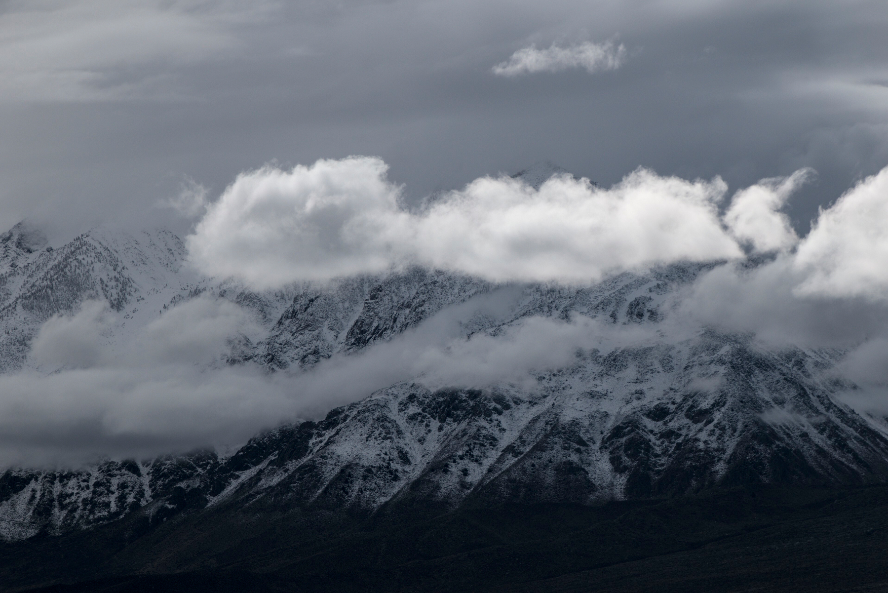 A high granite wall with fresh snow has long clouds floating along its face.