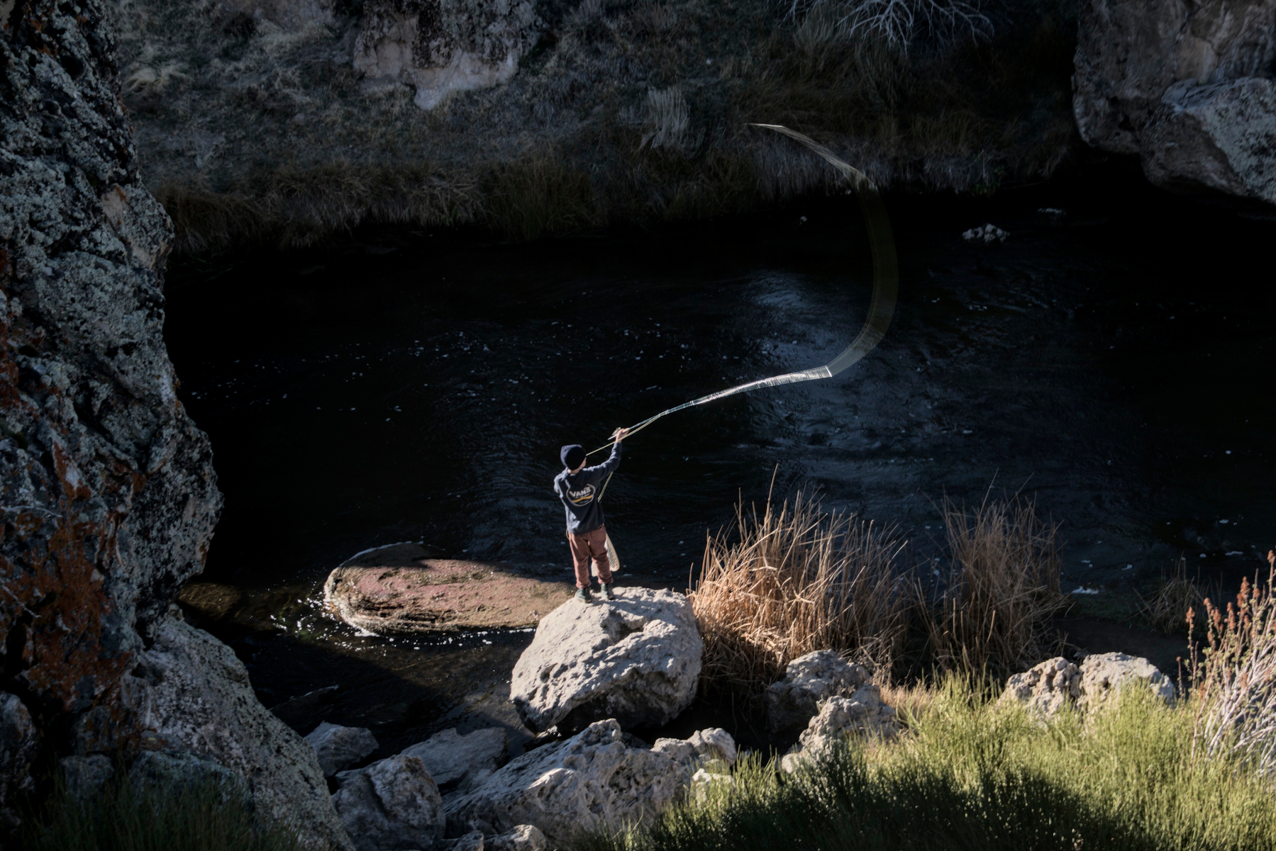 A teen stands on a rock at the edge of a river, casting a fishing line.